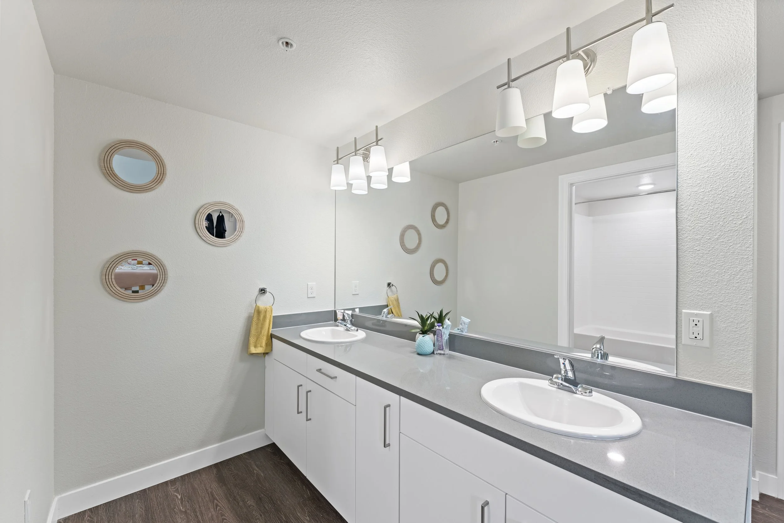 Bathroom vanity with two sinks, large mirror, white cabinets, modern light fixture, small plant, yellow towel, wall decorations, and a doorway leading to a bathtub/shower area.