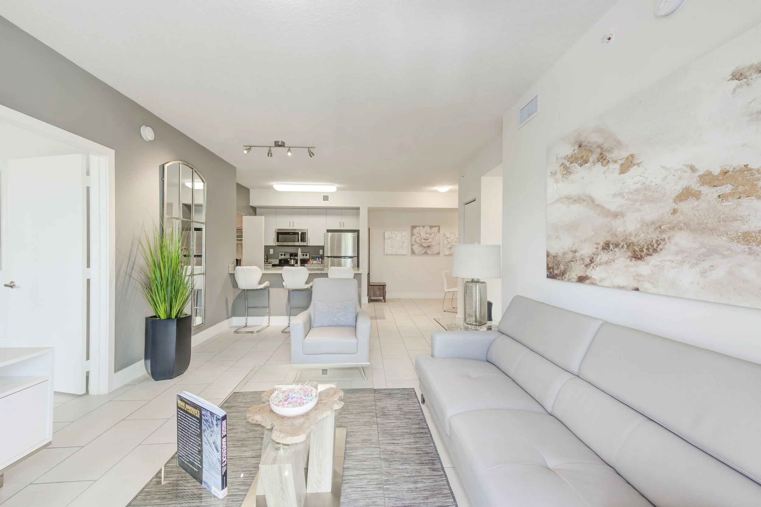 A modern, bright living room with a white and gray color scheme. There is a gray sofa, a white armchair, a glass coffee table with a decorative bowl, and a large abstract wall art. The background shows a kitchen with a breakfast bar, white chairs, an
