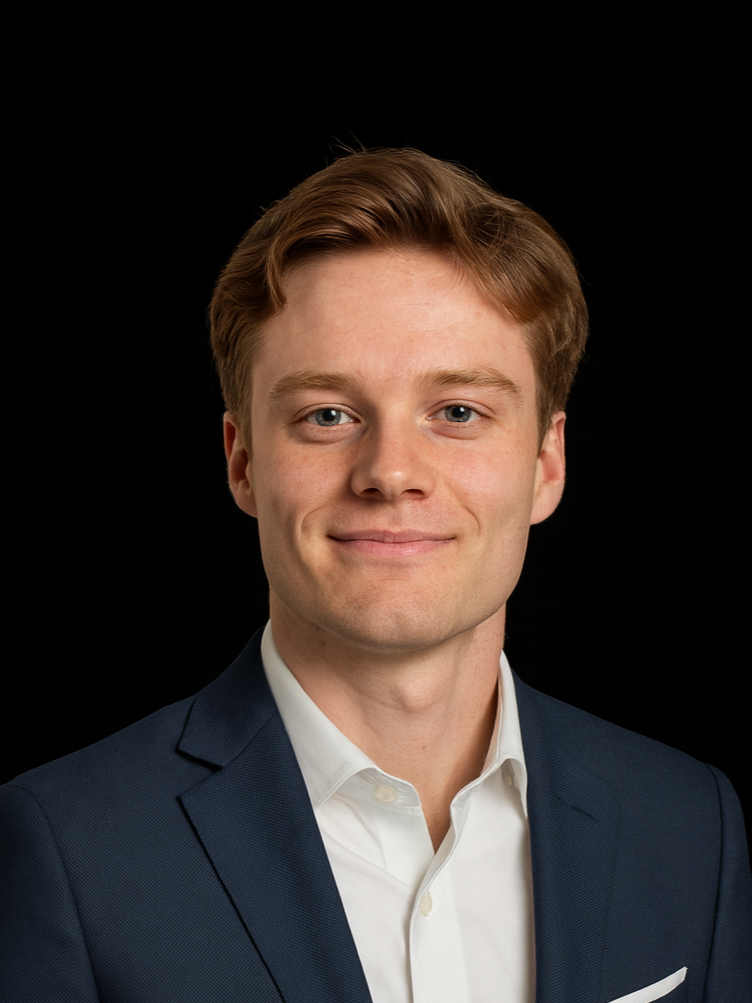 Portrait of a young man with short red hair, wearing a black suit, white shirt, and black tie, against a dark gray background.