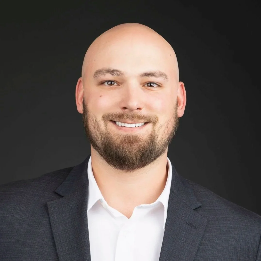 Headshot of a smiling man with a beard, wearing a dark suit jacket and white shirt, against a dark background.