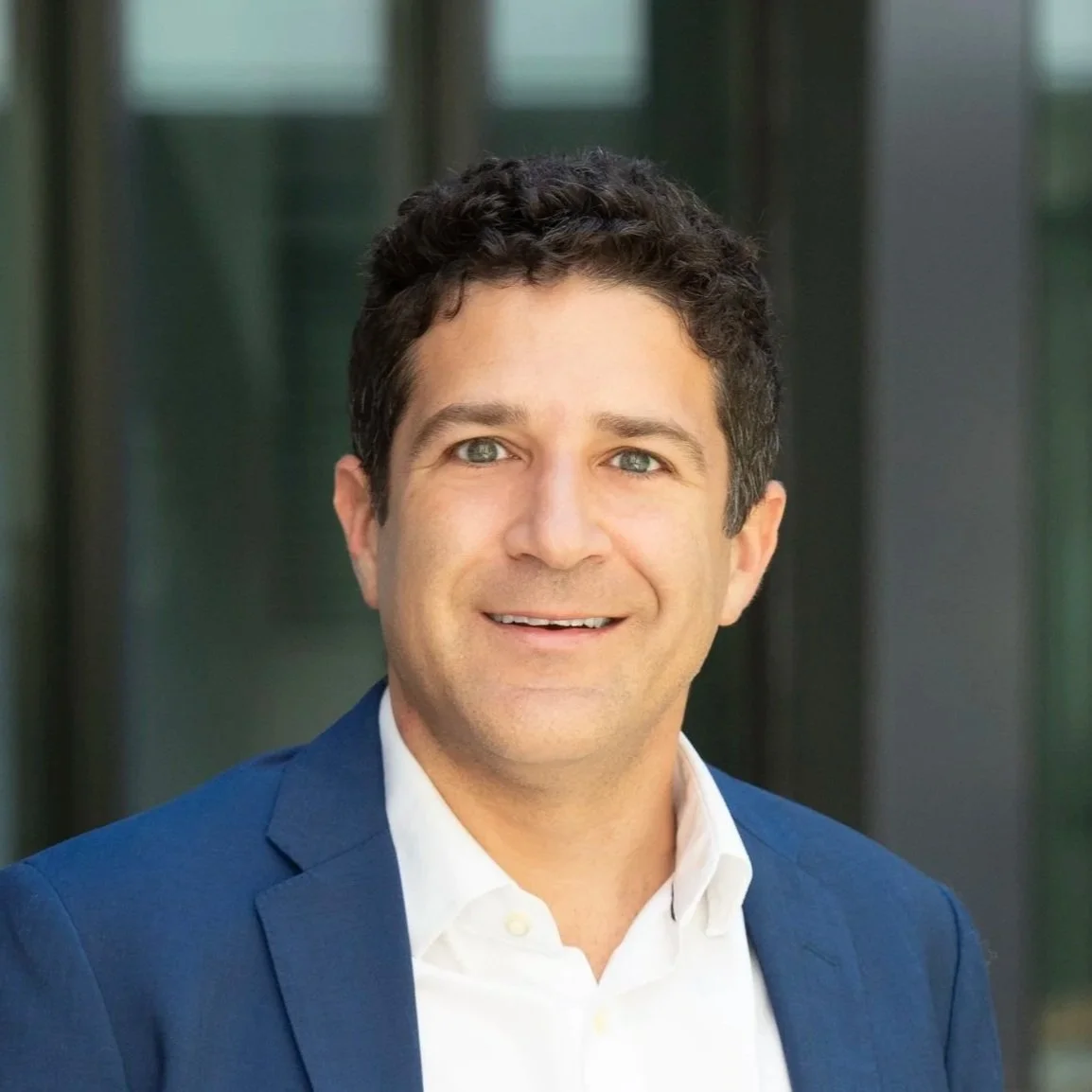 Headshot photo of a smiling man in a white shirt and blue blazer, standing outside with a modern glass building in the background.
