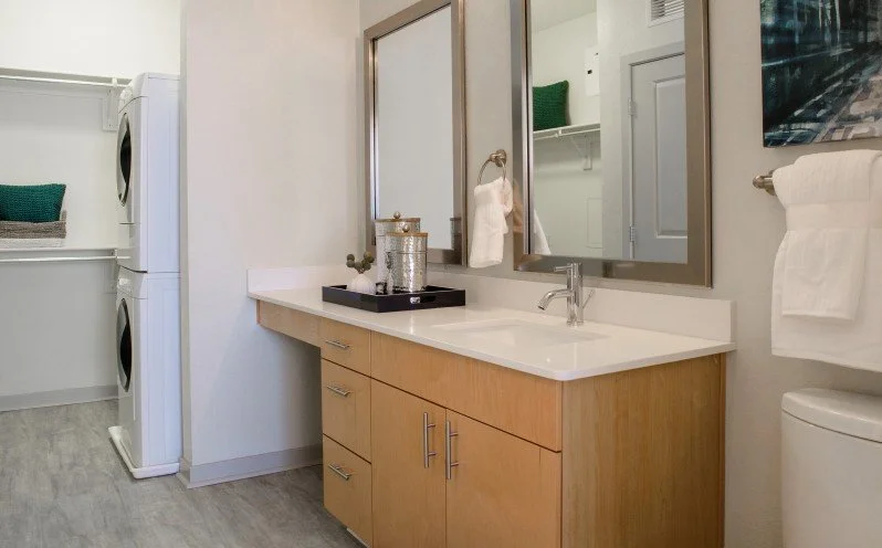 Bathroom vanity with a large mirror, faucet, and a tray with a decorative jar, next to a towel rack with a white towel. In the background, laundry machines and a laundry basket are visible.