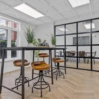 Modern office space with a high counter, four wooden bar stools, large windows, and potted plants.