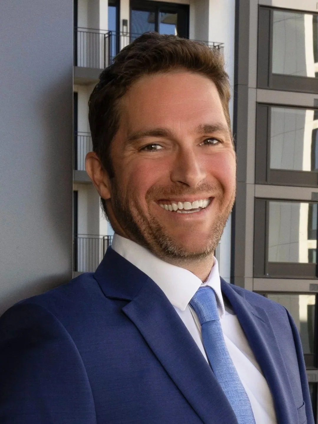 A smiling man in a blue suit and tie, standing outdoors in front of modern apartment buildings.