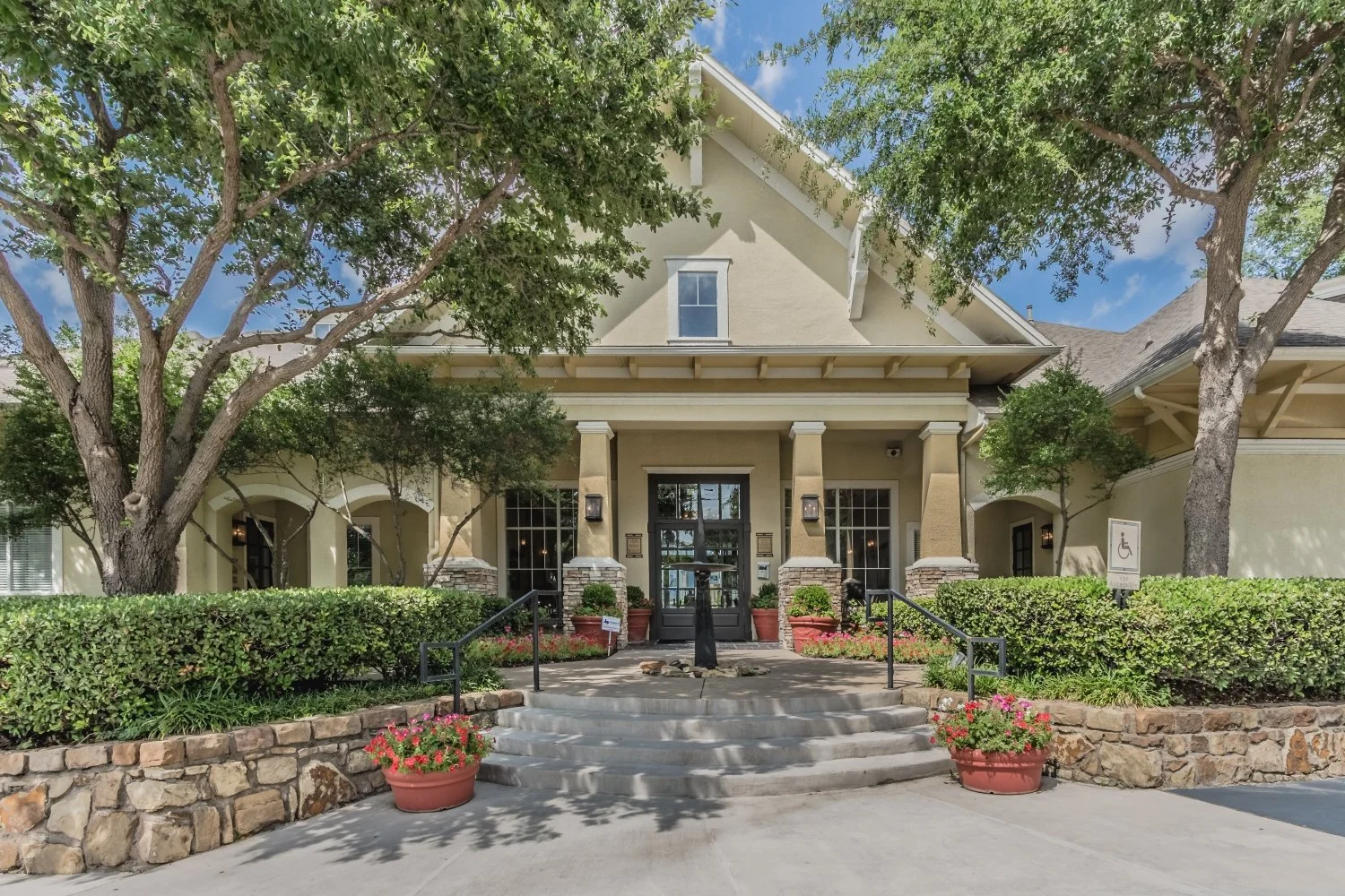 A large, beige residential house with a front porch and black door, surrounded by green trees and bushes, with a rock-lined stone wall and potted flowers near the entrance.