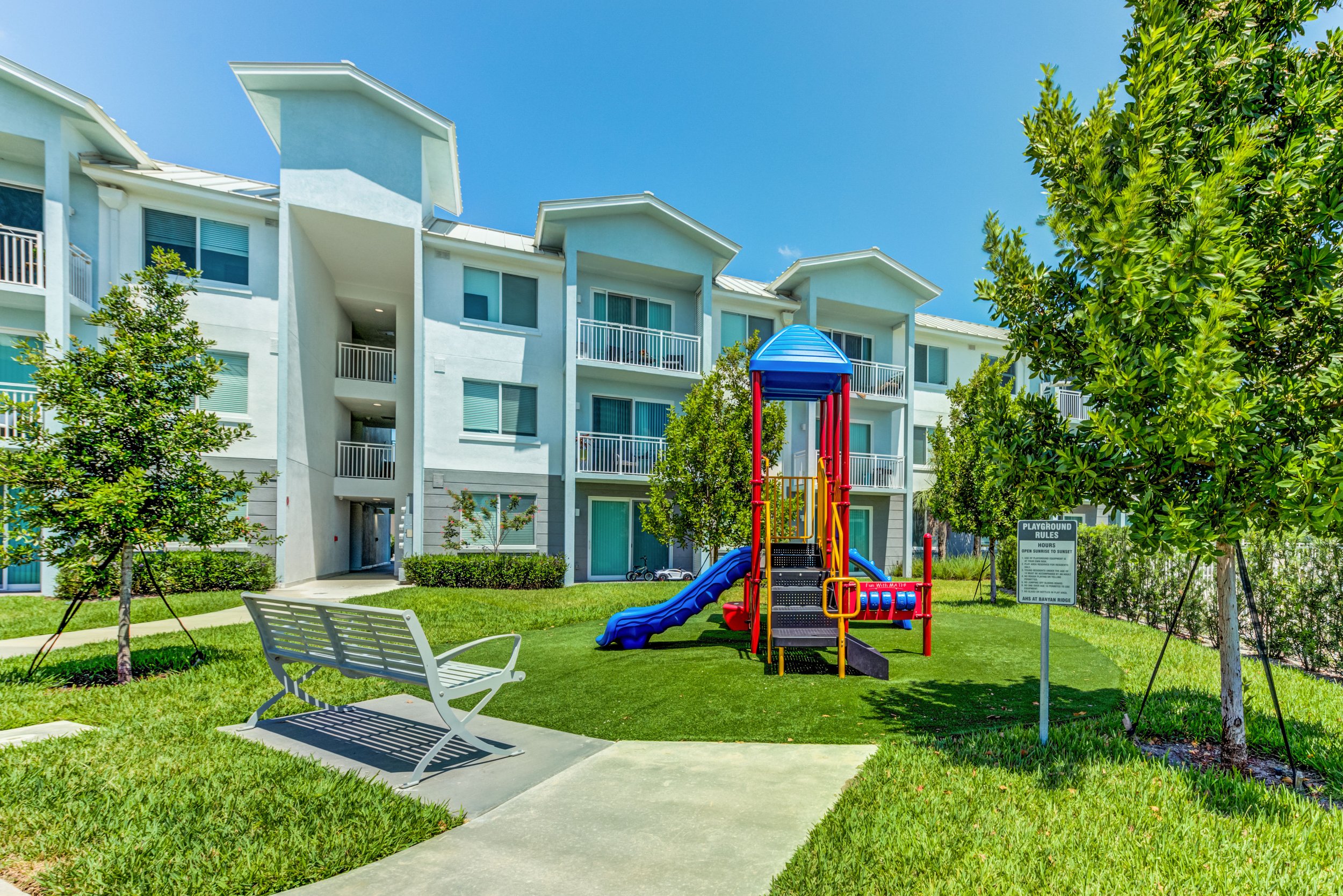 Children's playground with a slide and climbing structure in front of a modern white apartment building on a sunny day.