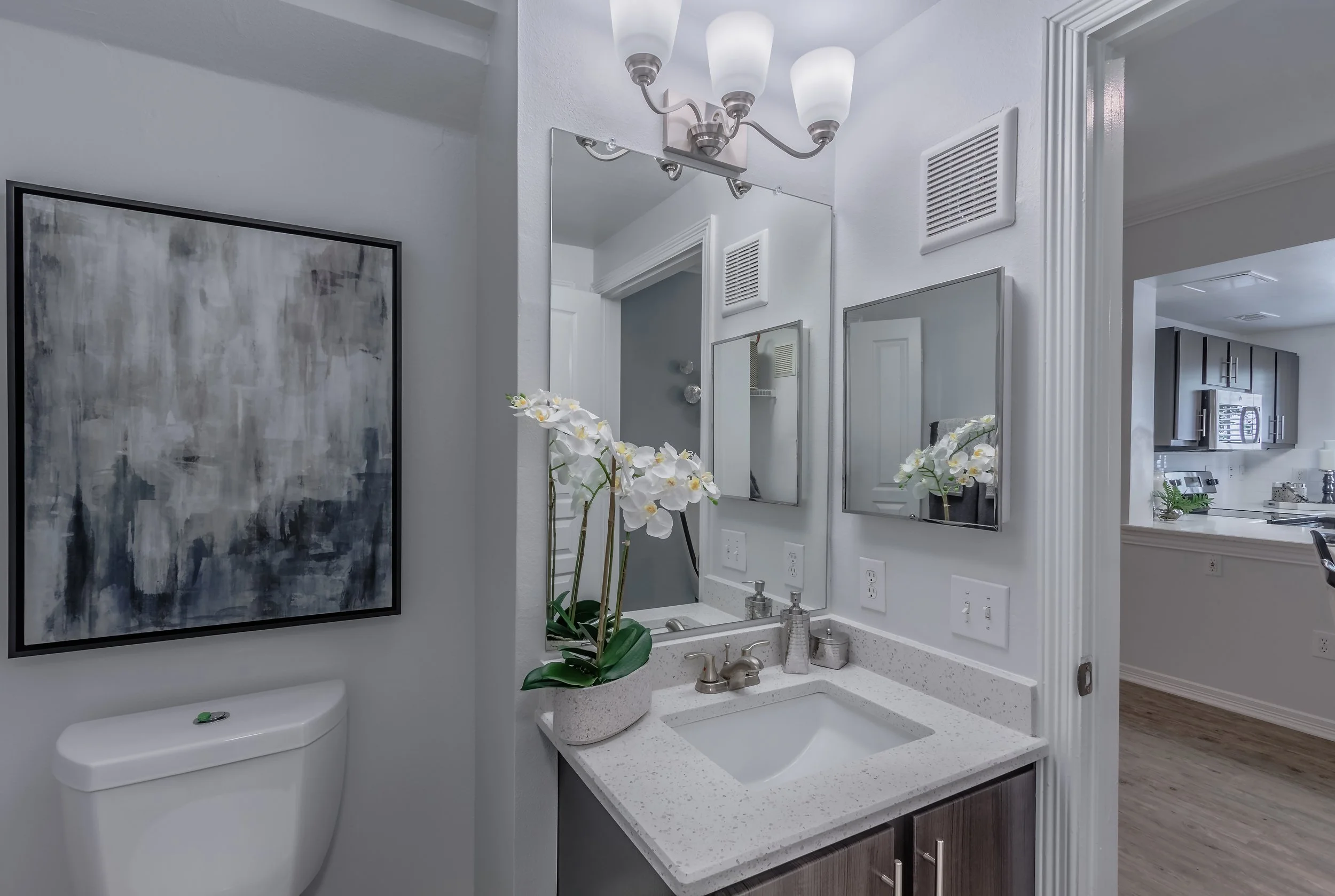 A modern bathroom vanity area with a white countertop, sink, and faucet. A potted white orchid decorates the counter. There are two small wall-mounted mirrors and a bottle of soap. The area is well-lit by a light fixture with five frosted glass shade