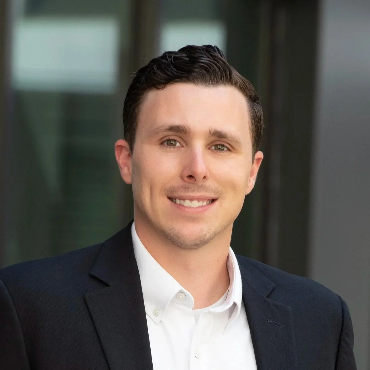 A portrait of a young man with dark hair, wearing a white shirt and dark blazer, smiling in front of a blurred background.