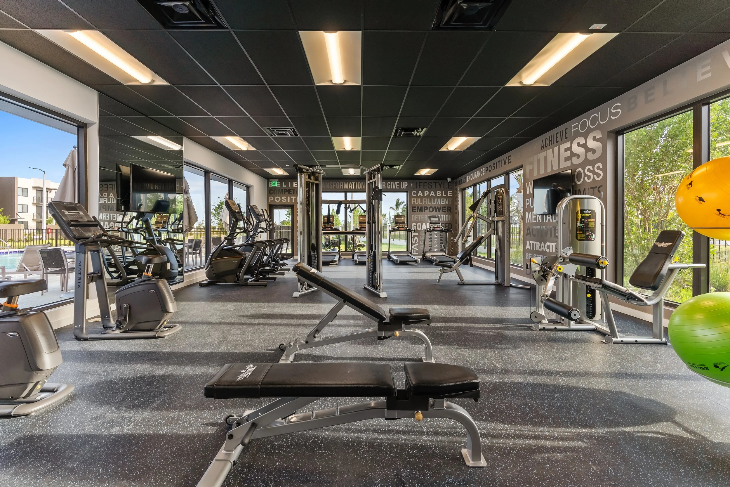 Empty gym with cardio machines, free weights, and workout equipment, with large windows showing an outdoor pool and green trees.