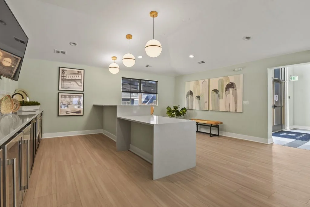 Interior of a modern, minimalistic kitchen and waiting area with light green walls, wooden flooring, a kitchen counter, wall art, and pendant lights.