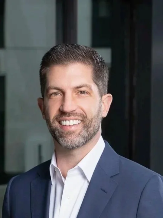 A smiling man with slicked-back hair, a beard, and a checkered blazer standing outside in front of a multi-story apartment building with balconies and streetlights.