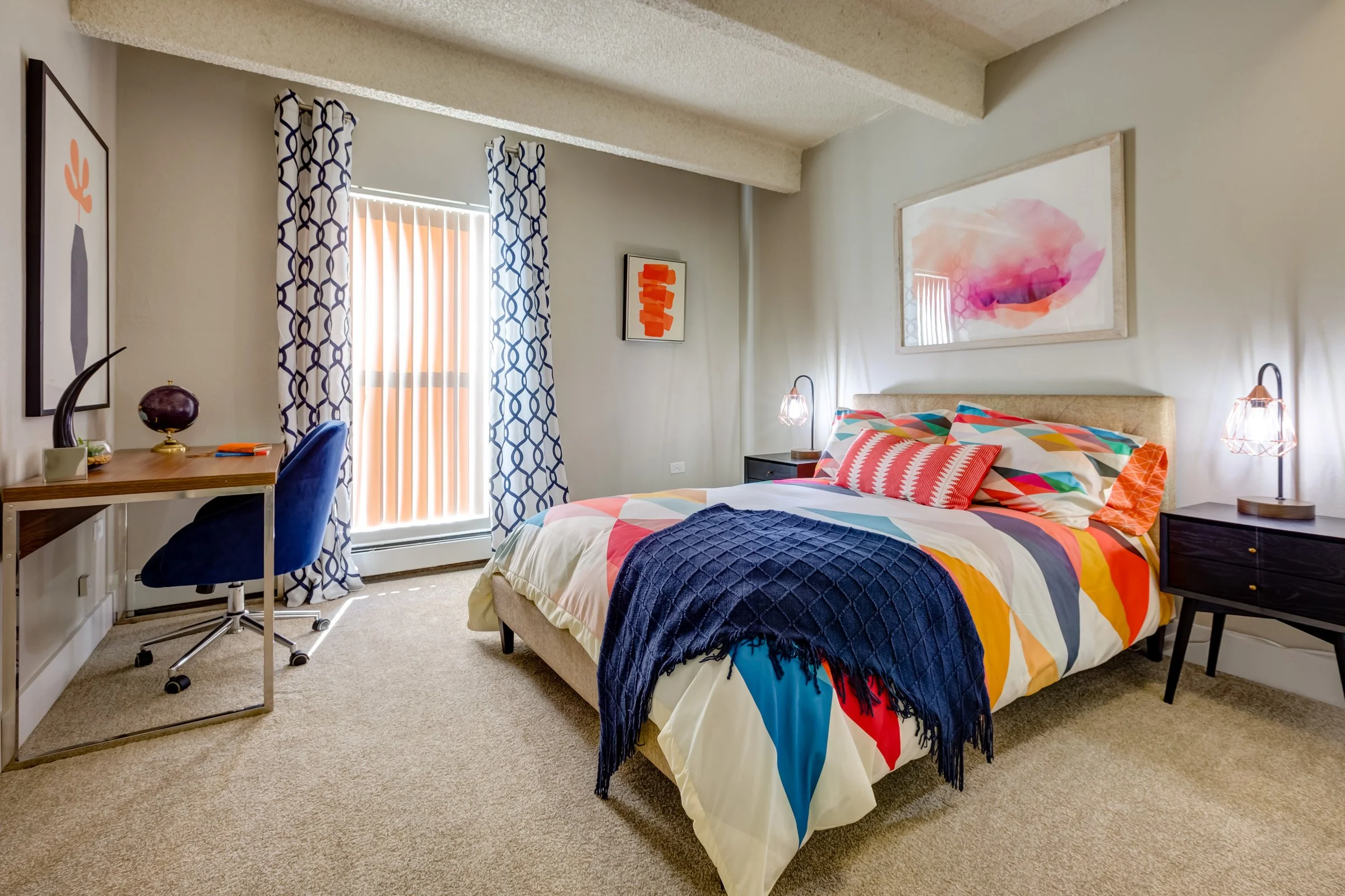 Bright bedroom with a bed featuring colorful geometric-patterned bedding, dark nightstands with lamps on each side, a desk with a blue chair, and framed art on the walls. There are curtains with a blue pattern and vertical blinds covering a window.