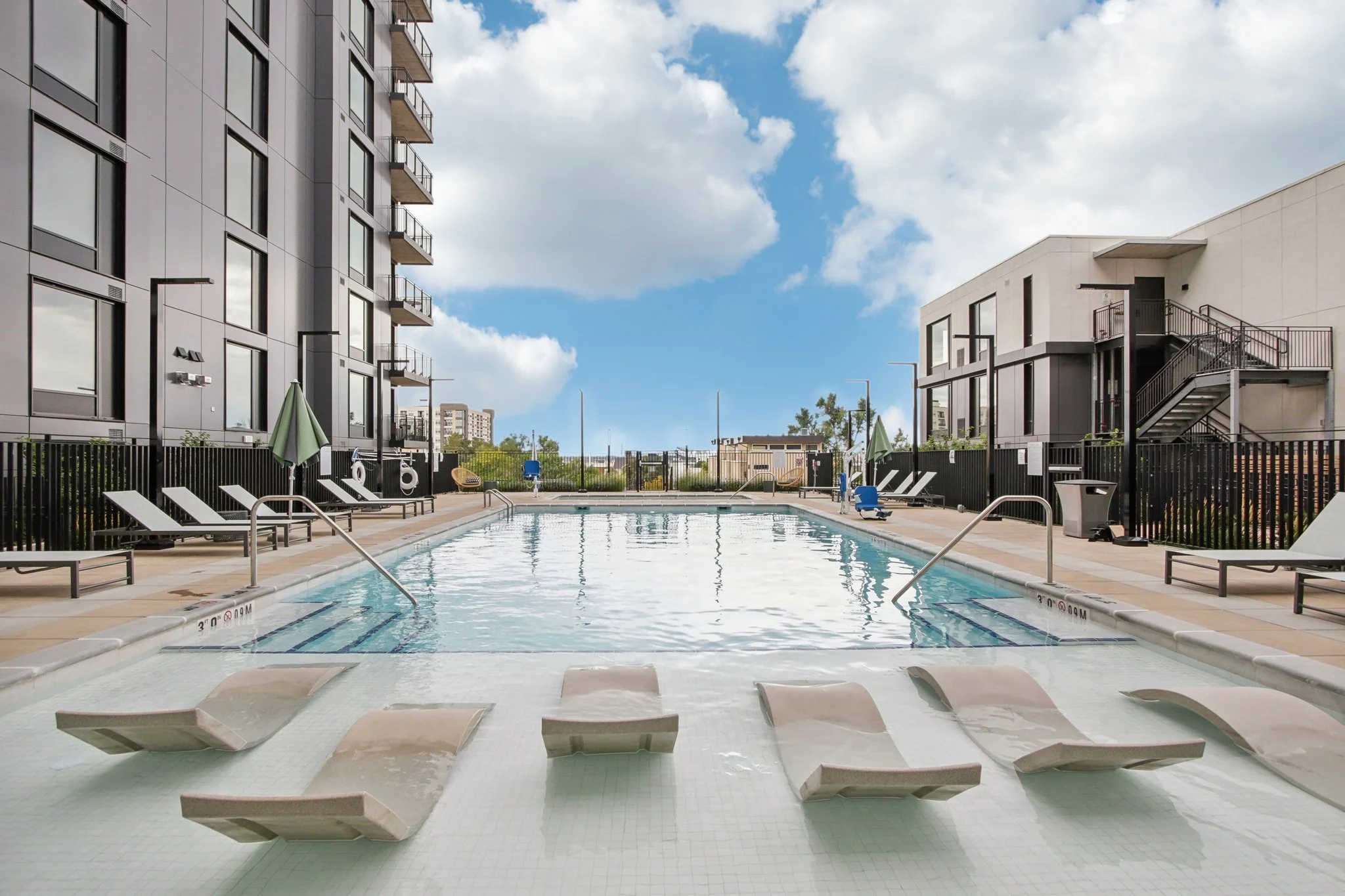 Empty outdoor swimming pool at a modern apartment complex with lounge chairs, umbrellas, and surrounding buildings under a partly cloudy sky.