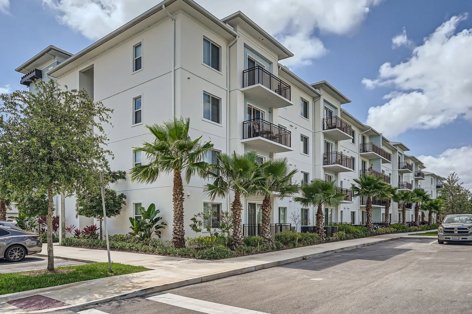 A modern white apartment building with multiple floors and balconies, lined with palm trees and parked cars in the foreground under a partly cloudy sky.