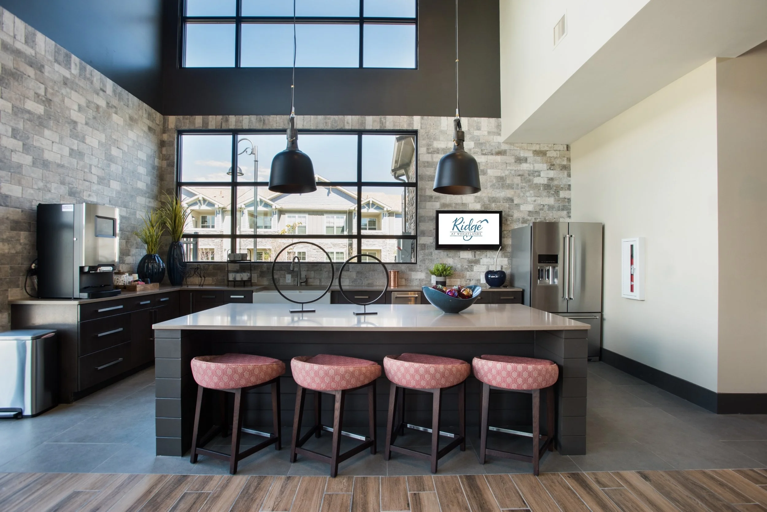 Modern kitchen with a large island, four pink upholstered barstools, black pendant lights, stainless steel refrigerator, window with views of outside buildings, and decorative vases and bowl on the countertops.