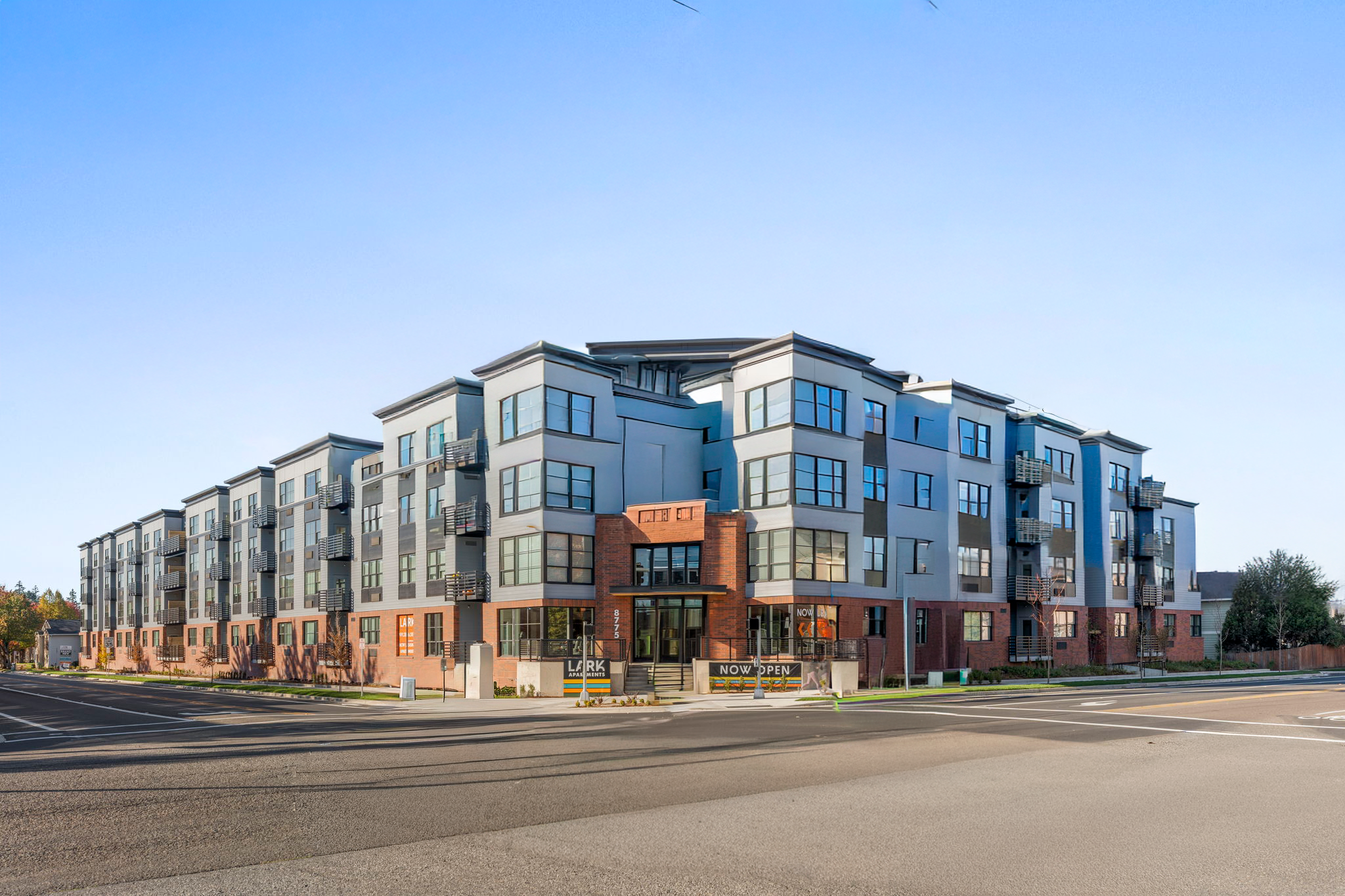 Modern multi-story apartment building with large windows and small balconies, located on a city street with a clear blue sky.