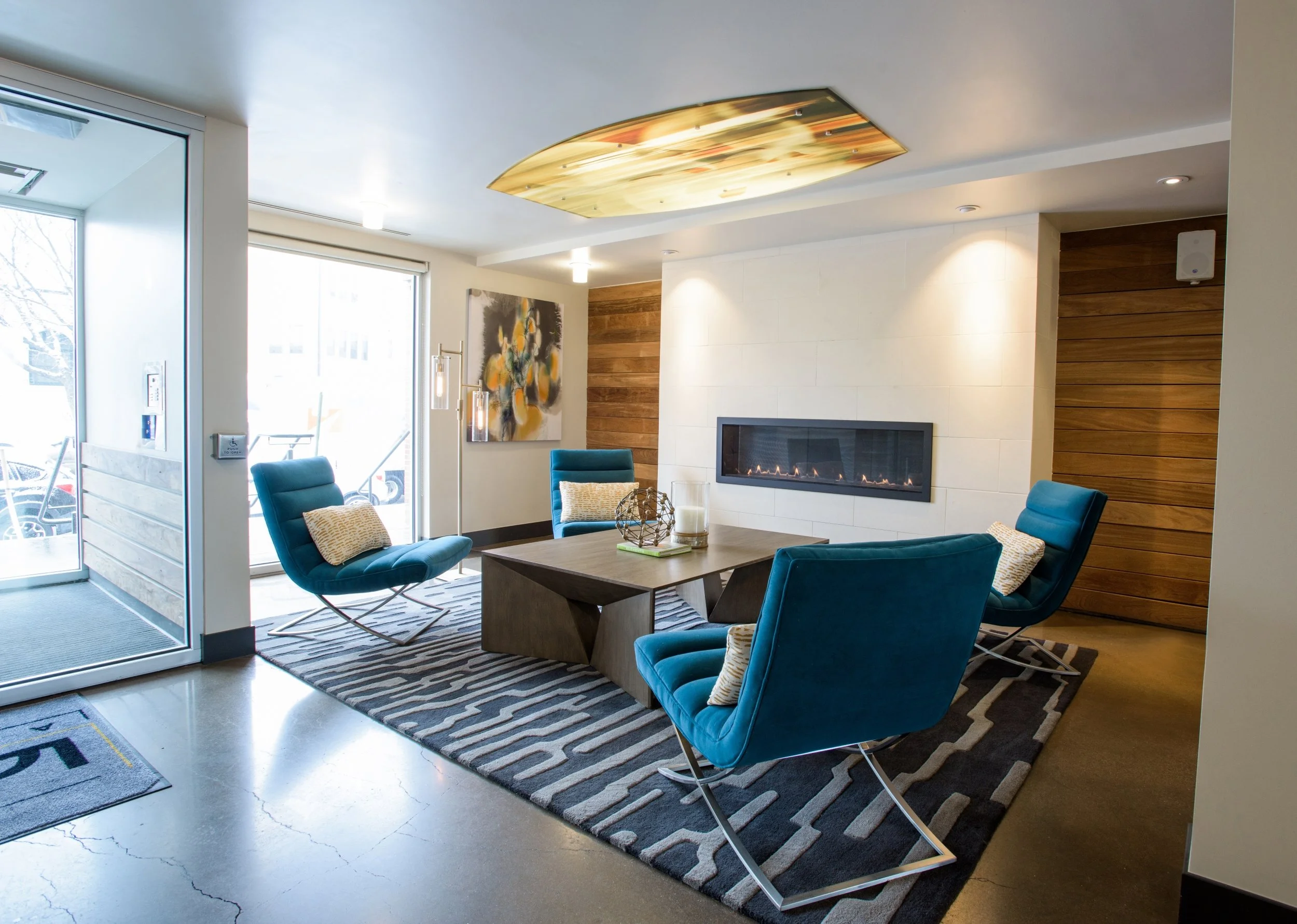 Modern lounge area with blue chairs, a wooden coffee table, a fireplace, abstract artwork, and large windows letting in natural light.