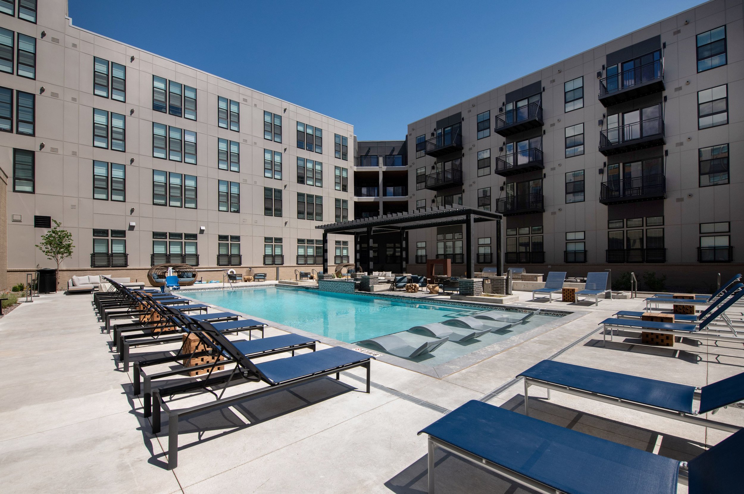 Outdoor heated pool with lounge chairs, shade structures, and water features, surrounded by a modern apartment complex under a clear blue sky.