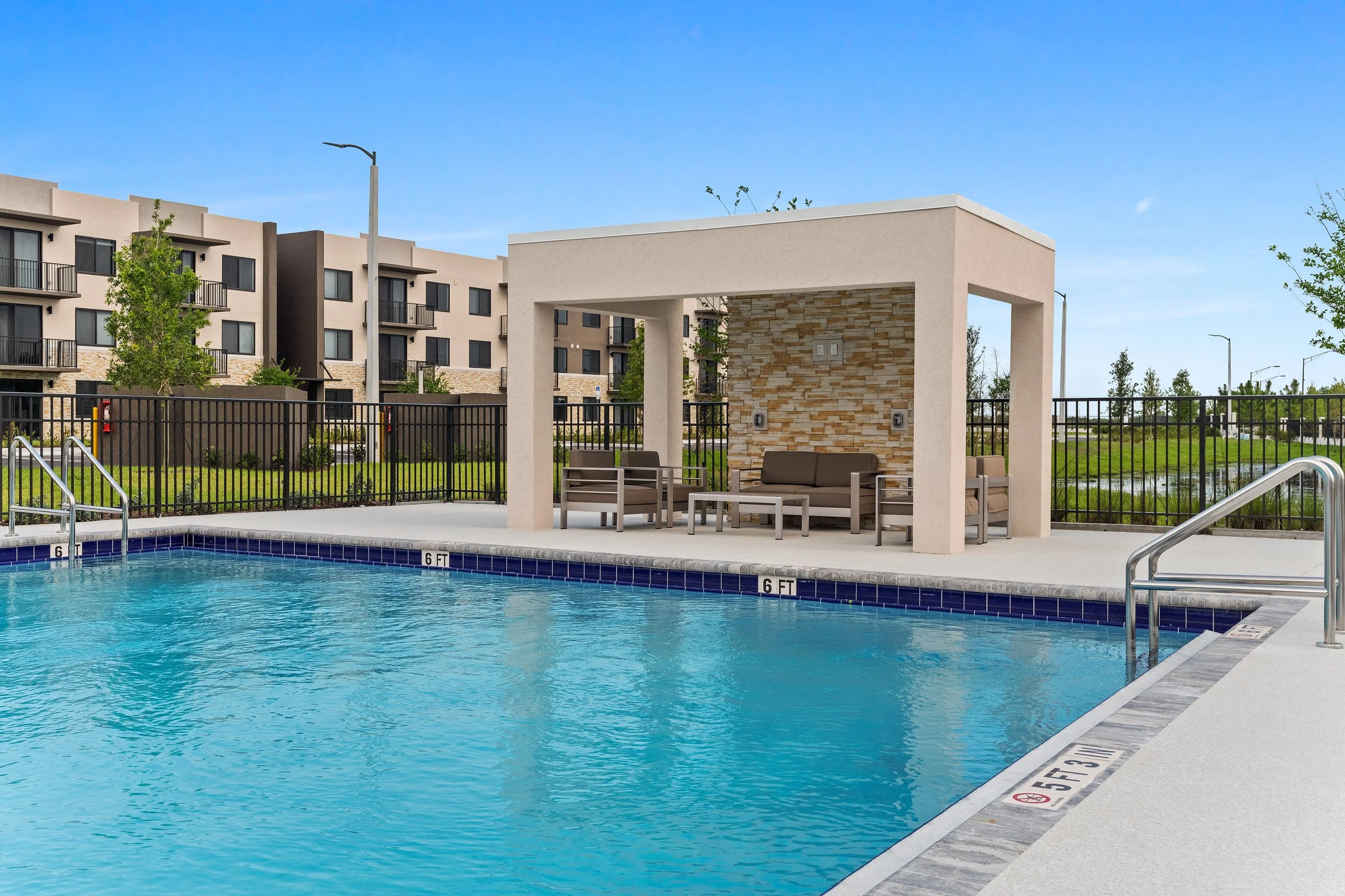 Swimming pool with a shaded seating area and chairs, surrounded by a black fence, in an apartment complex with modern buildings in the background under a blue sky.