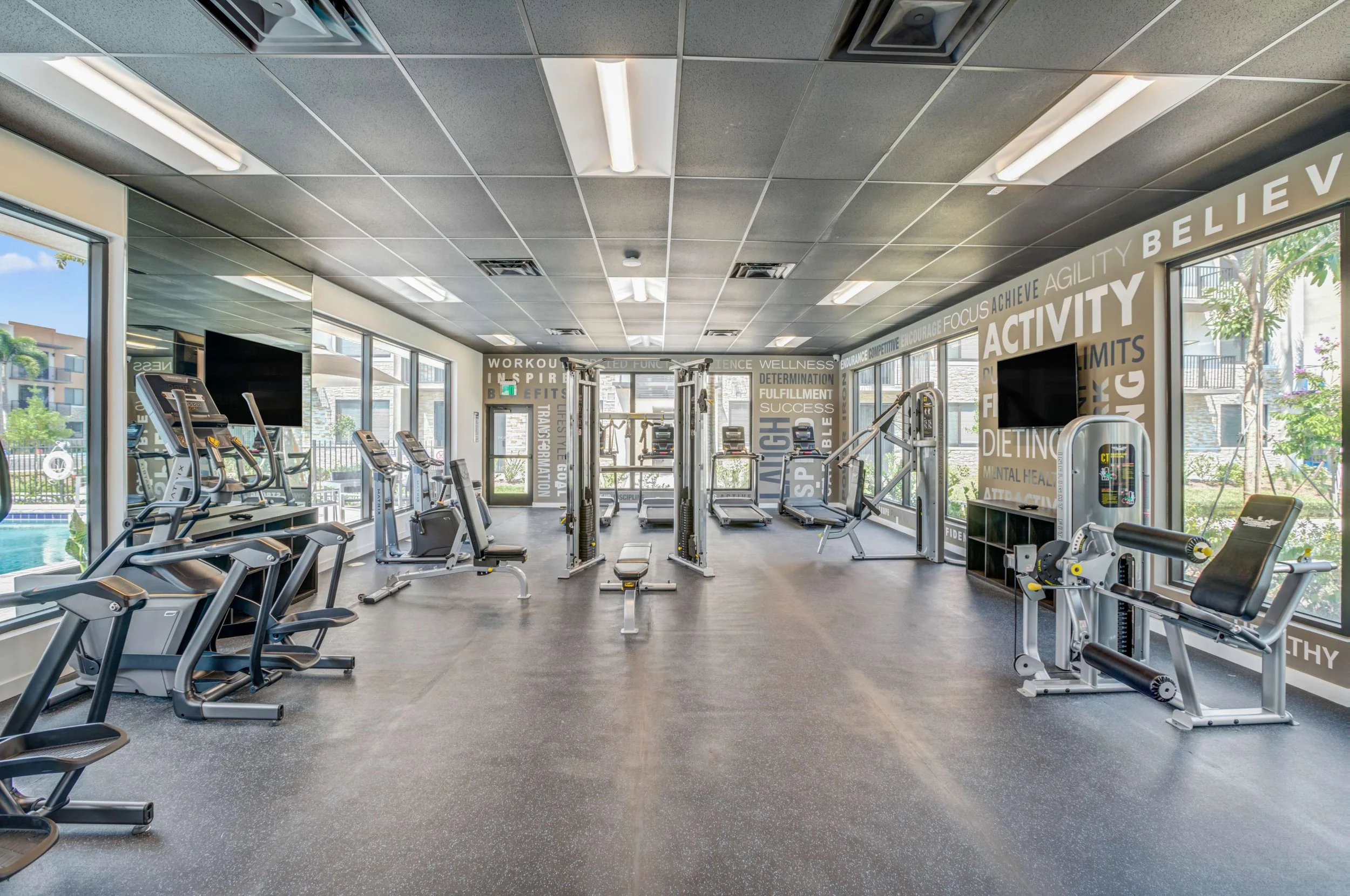 A modern gym with various exercise equipment, including treadmills and weight machines, in a bright space with large windows and motivational words on the walls.