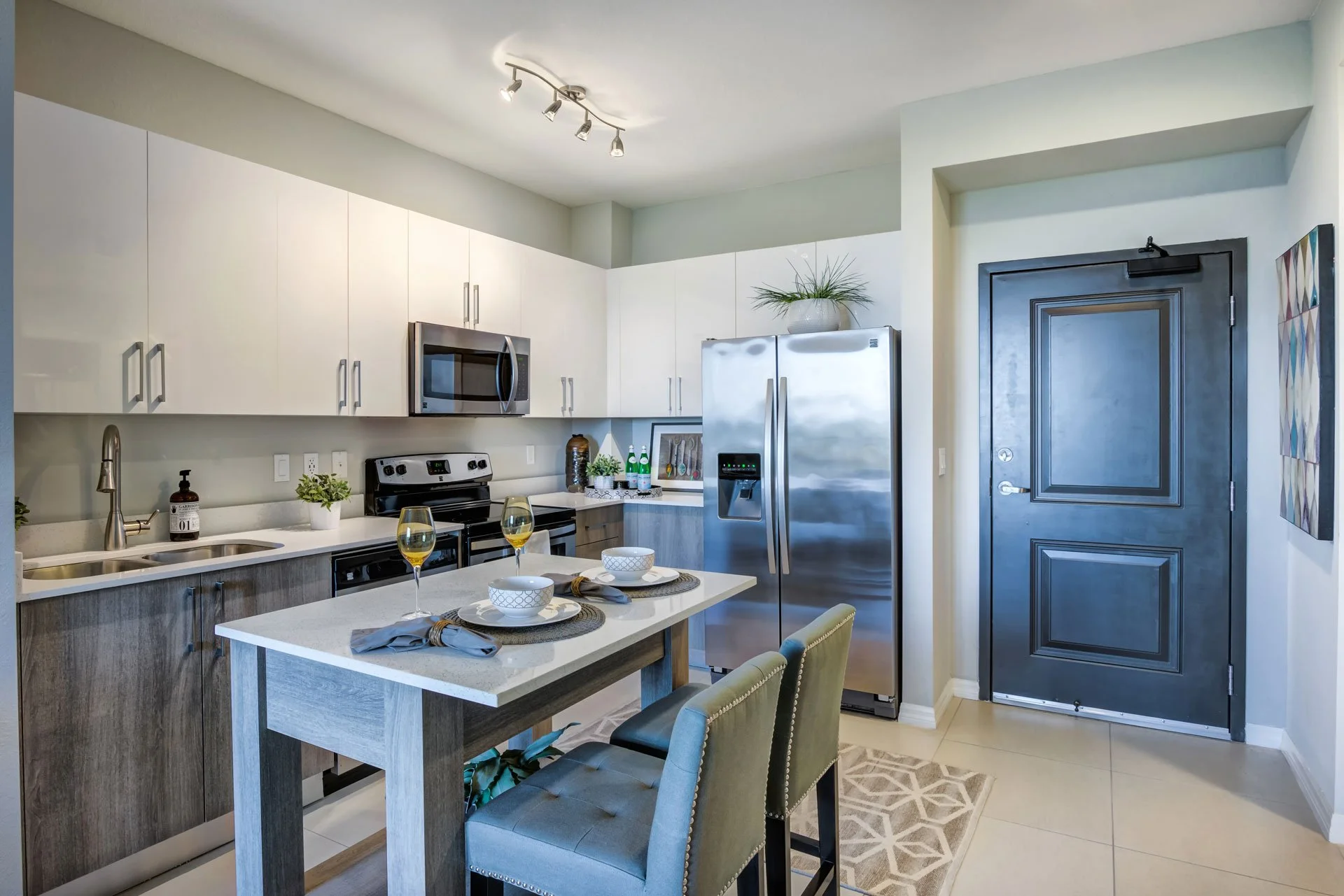 Modern kitchen with white upper cabinets, wood lower cabinets, stainless steel appliances including fridge, microwave, and oven. A small dining table with two chairs, set with plates, bowls, napkins, and glasses of white wine. Potted plants and decor