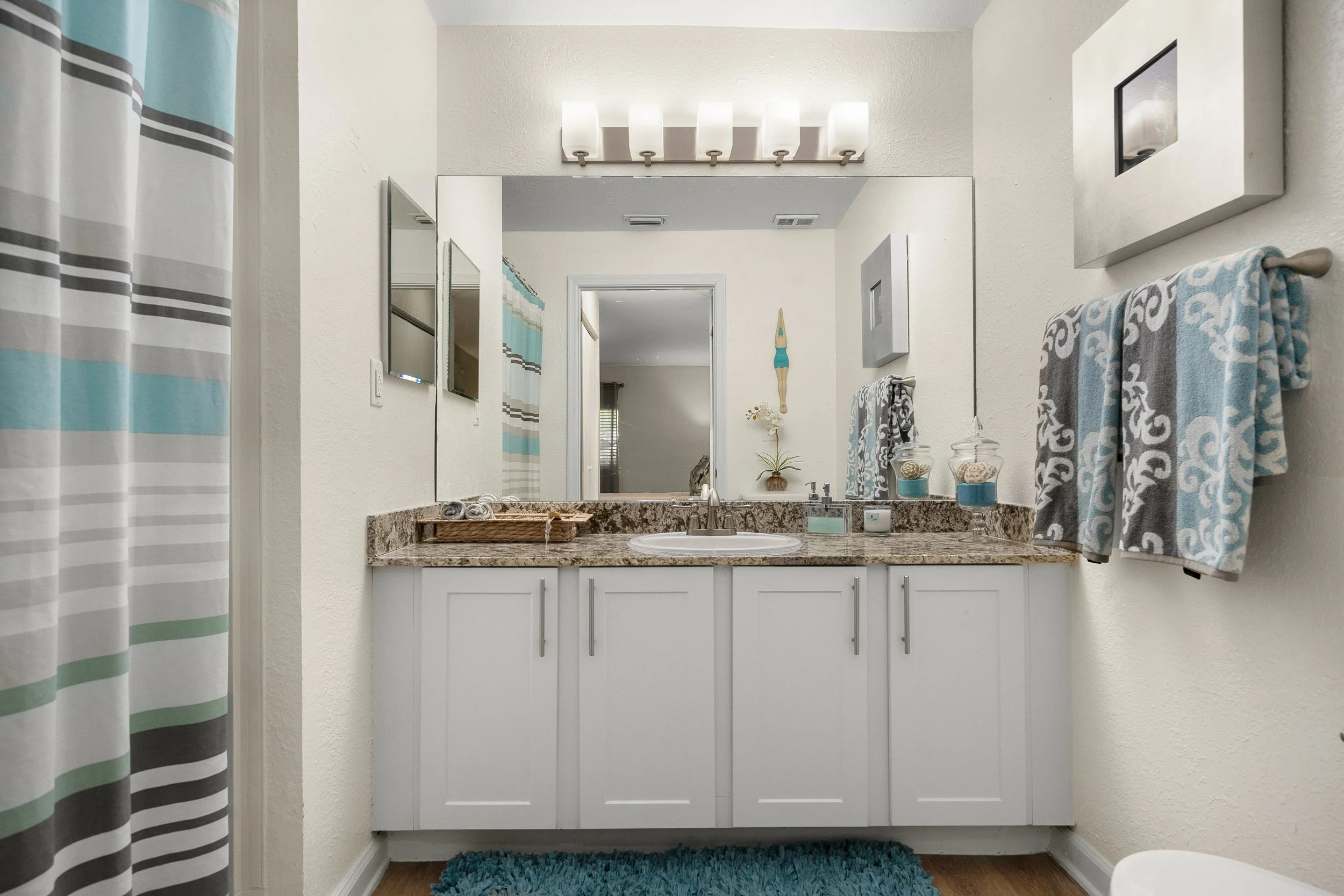 Bathroom with a white vanity, granite countertop, large mirror, wall-mounted light fixture, two small mirrors, towels hanging on a towel bar, a small potted plant, decorative jars, and a blue bath mat.