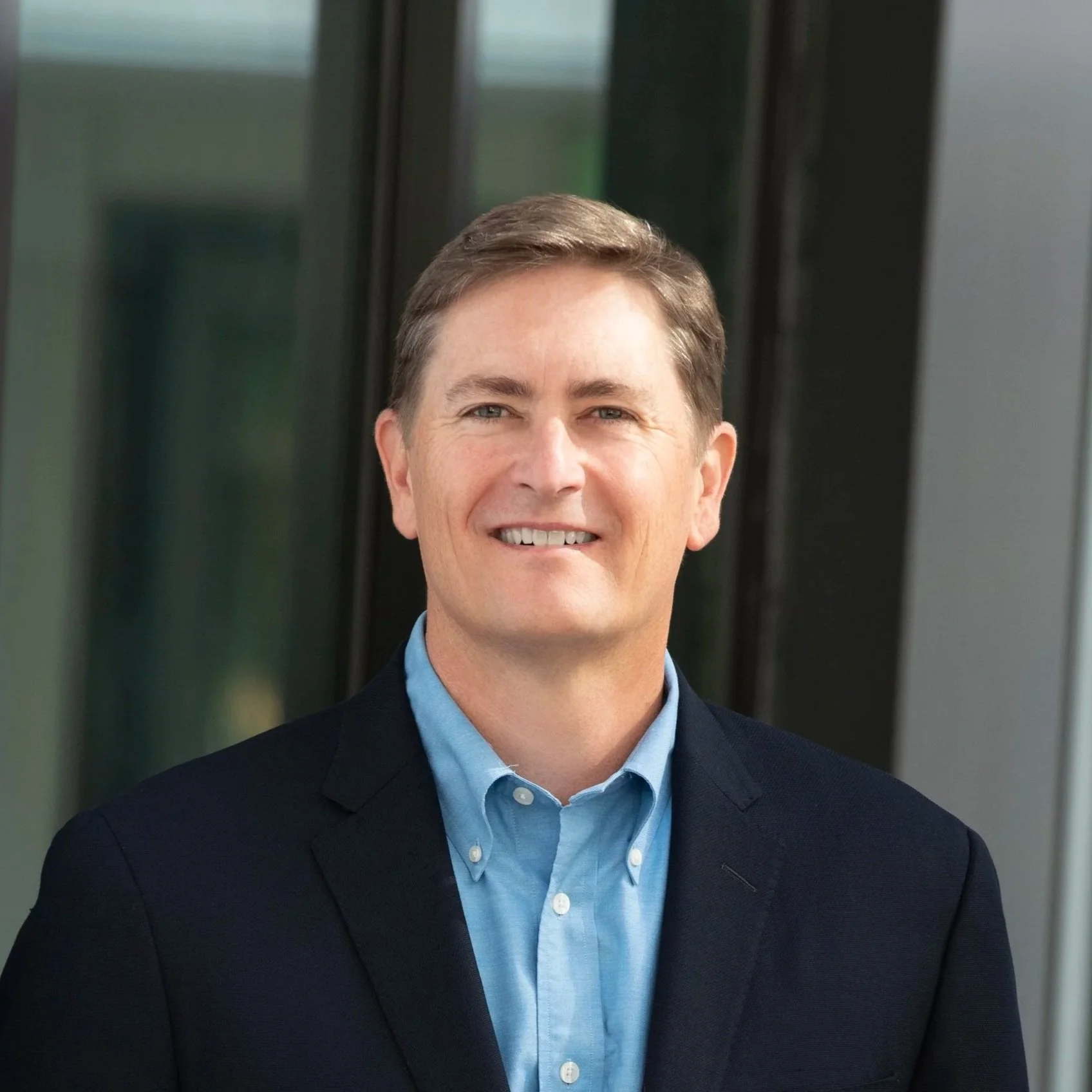 Professional portrait of a middle-aged man in a navy blazer and light blue shirt, standing outside near a modern building with glass windows.
