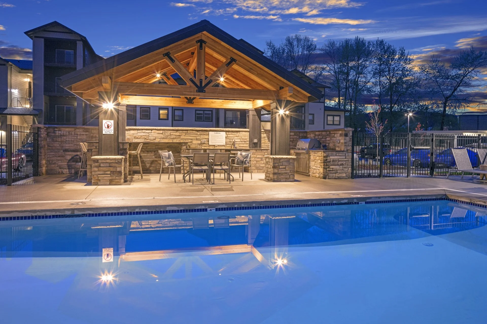 An outdoor pool area at sunset with a covered seating area featuring table and chairs, surrounded by a fence and residential buildings in the background.