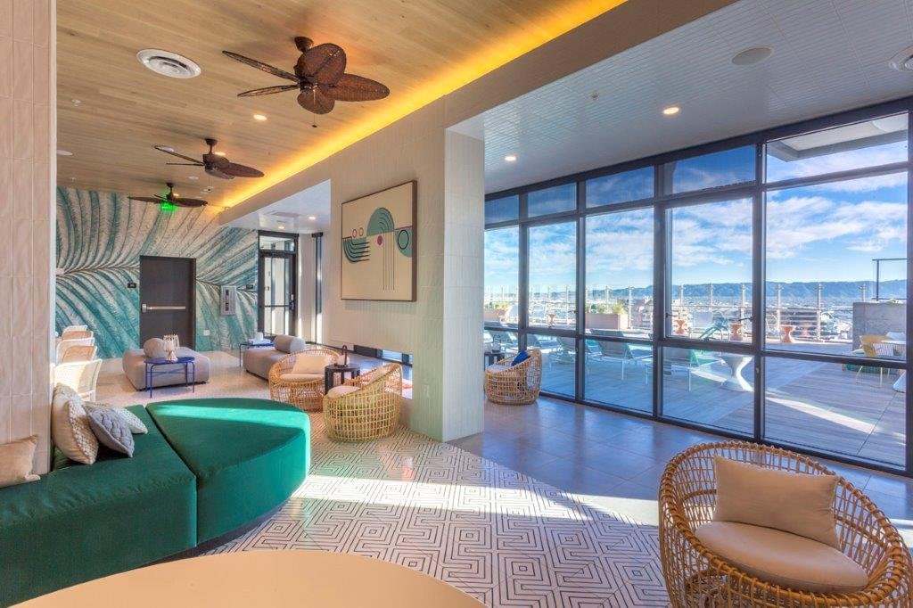 A modern lounge with large windows overlooking a marina, featuring wicker chairs, ceiling fans, and colorful abstract art.