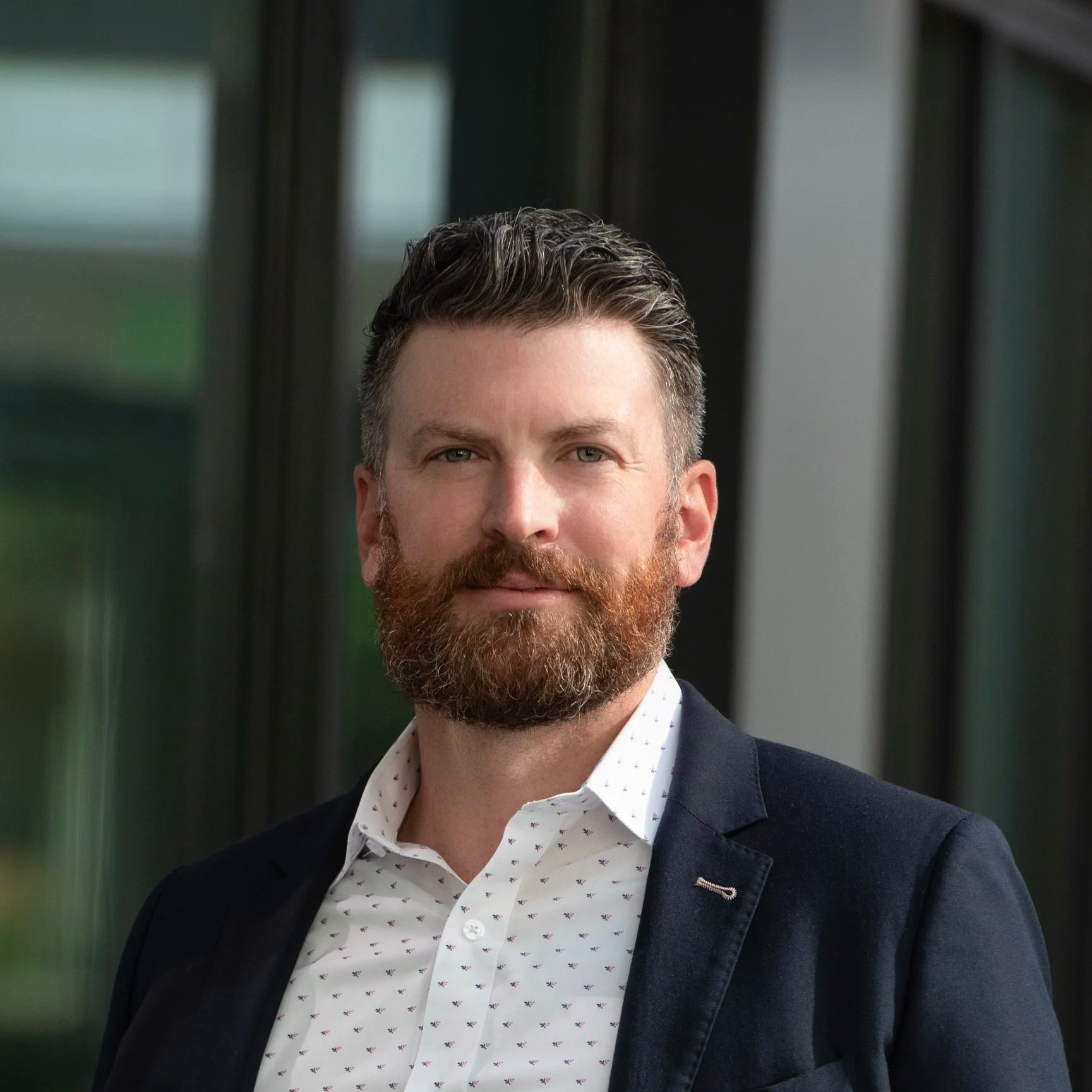 A man with a beard and styled hair wearing a white patterned shirt and dark blazer, standing indoors near large windows.