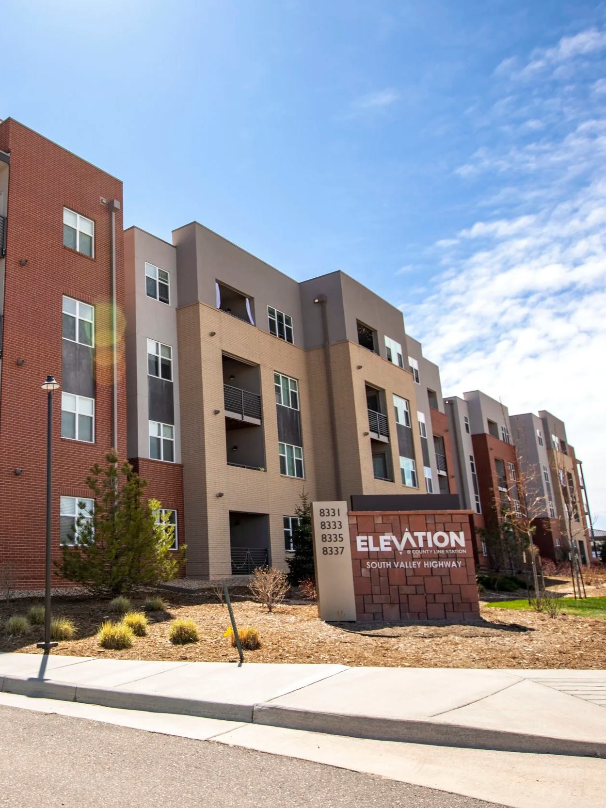 A modern multi-story apartment building with a sign reading 'Elevation, South Valley Highway', and addresses 8331 to 8337, on a sunny day with a clear blue sky.