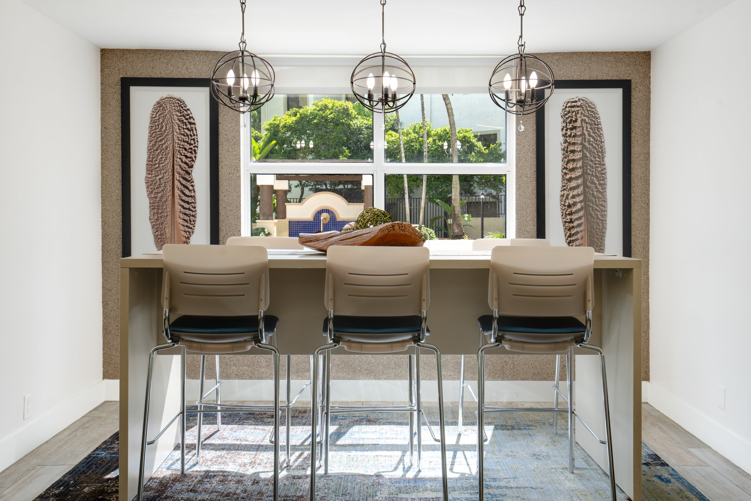 Modern dining area with a bar-height table and three beige chairs facing a large window. The window reveals a lush outdoor garden with trees and a fountain. The wall behind the window has two framed art pieces of zebra stripes, and three spherical pe