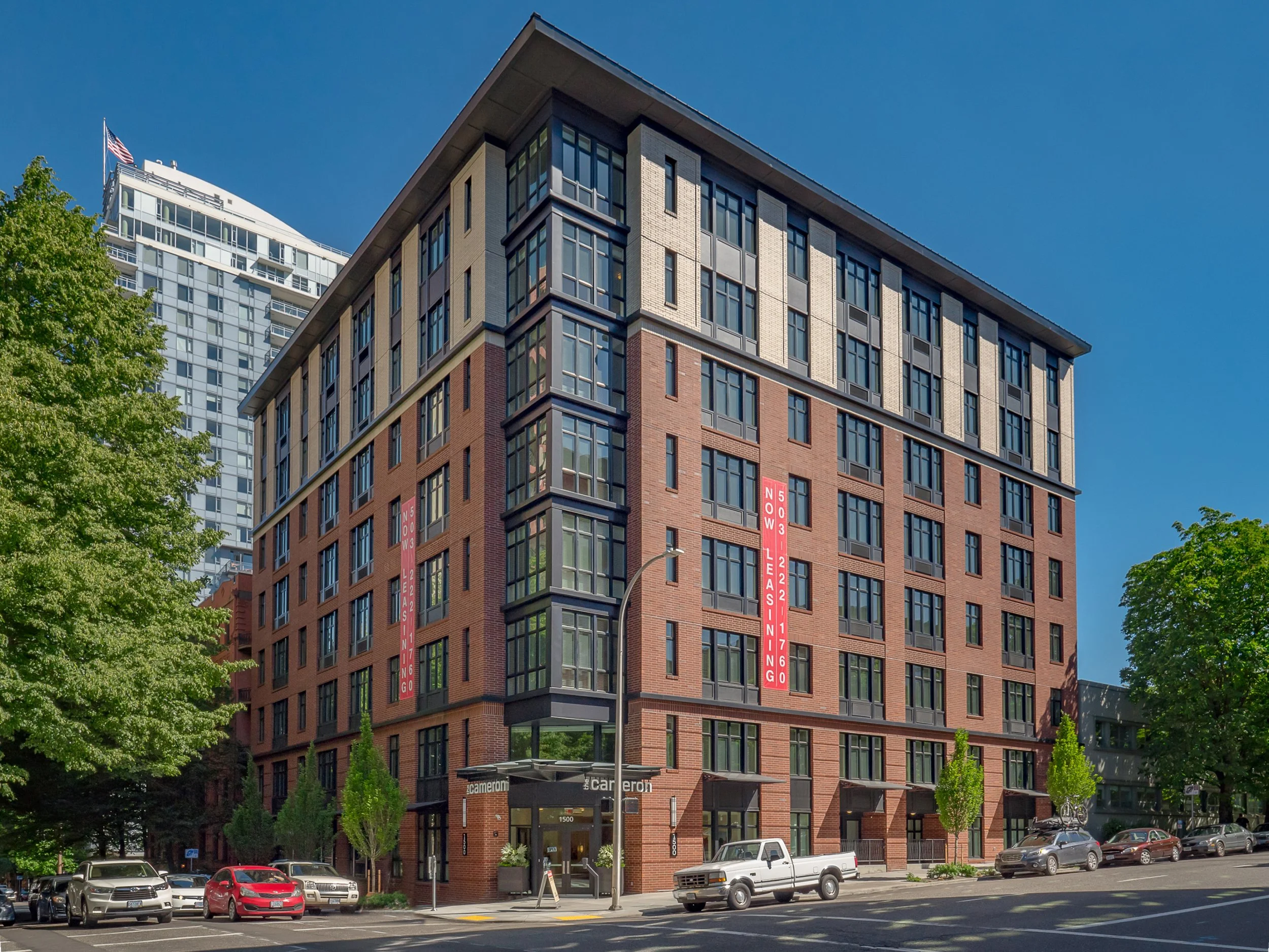 A multi-story modern apartment building with brick and glass exterior, surrounded by trees and parked cars, under a clear blue sky. There are vertical red banners on the building indicating leasing information.