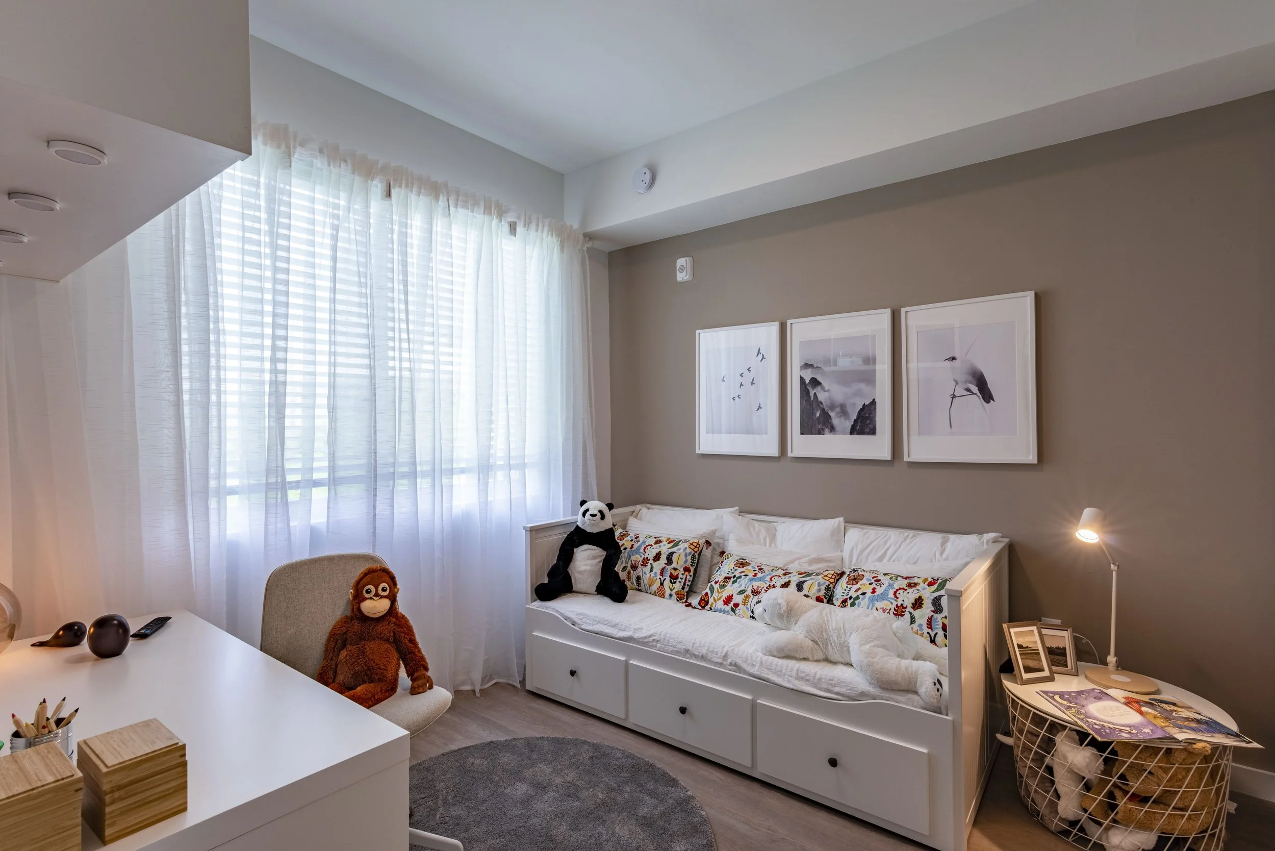 A cozy bedroom with a white daybed, decorated with plush toys and colorful pillows, and a round gray rug. The wall features three framed black-and-white prints of birds and nature.