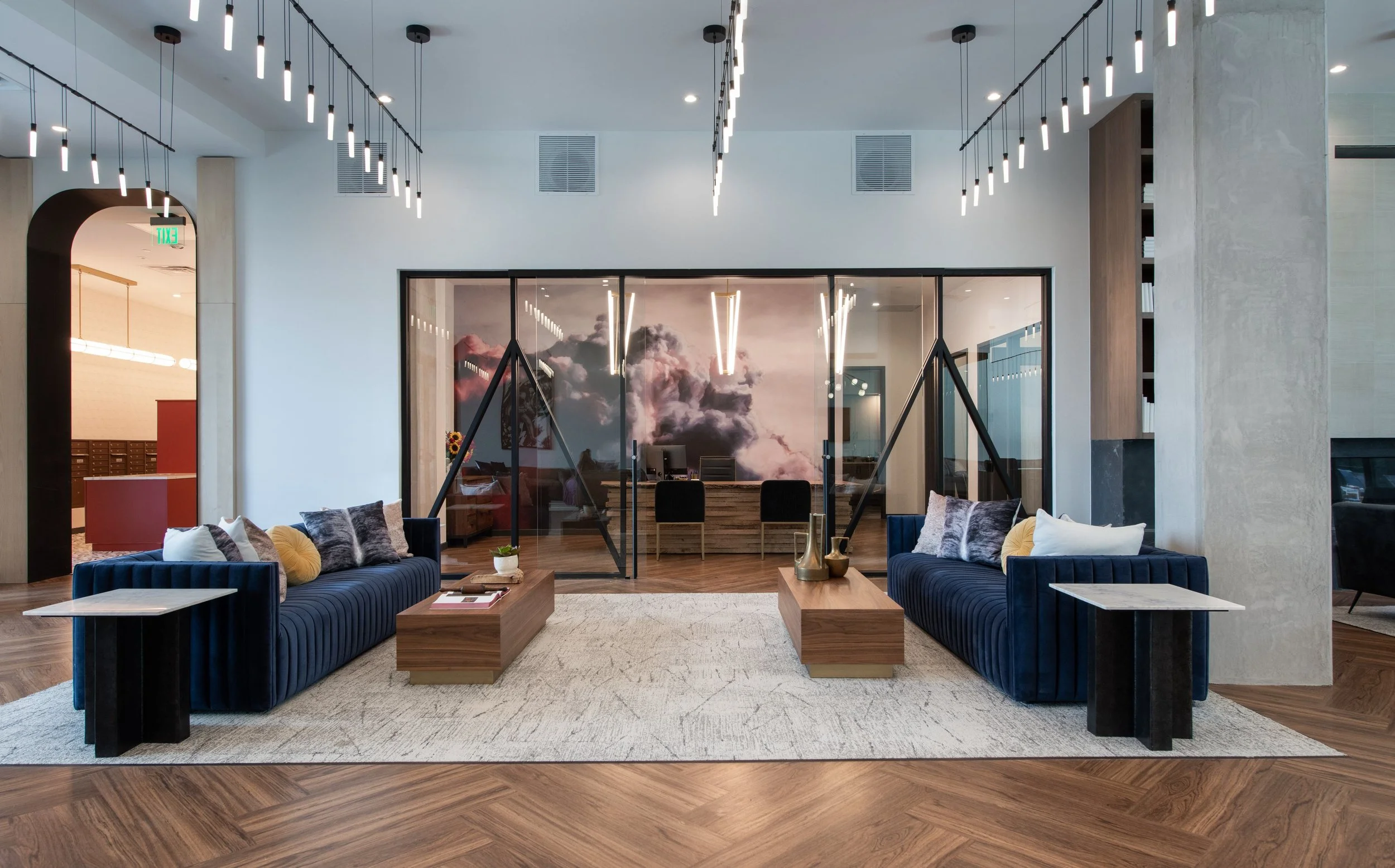 Modern hotel lobby with two navy velvet sofas on either side of a cream rug, wooden coffee tables, and throw pillows. Glass walls reveal a skylit office area with desks and chairs, and scenic cloud mural.