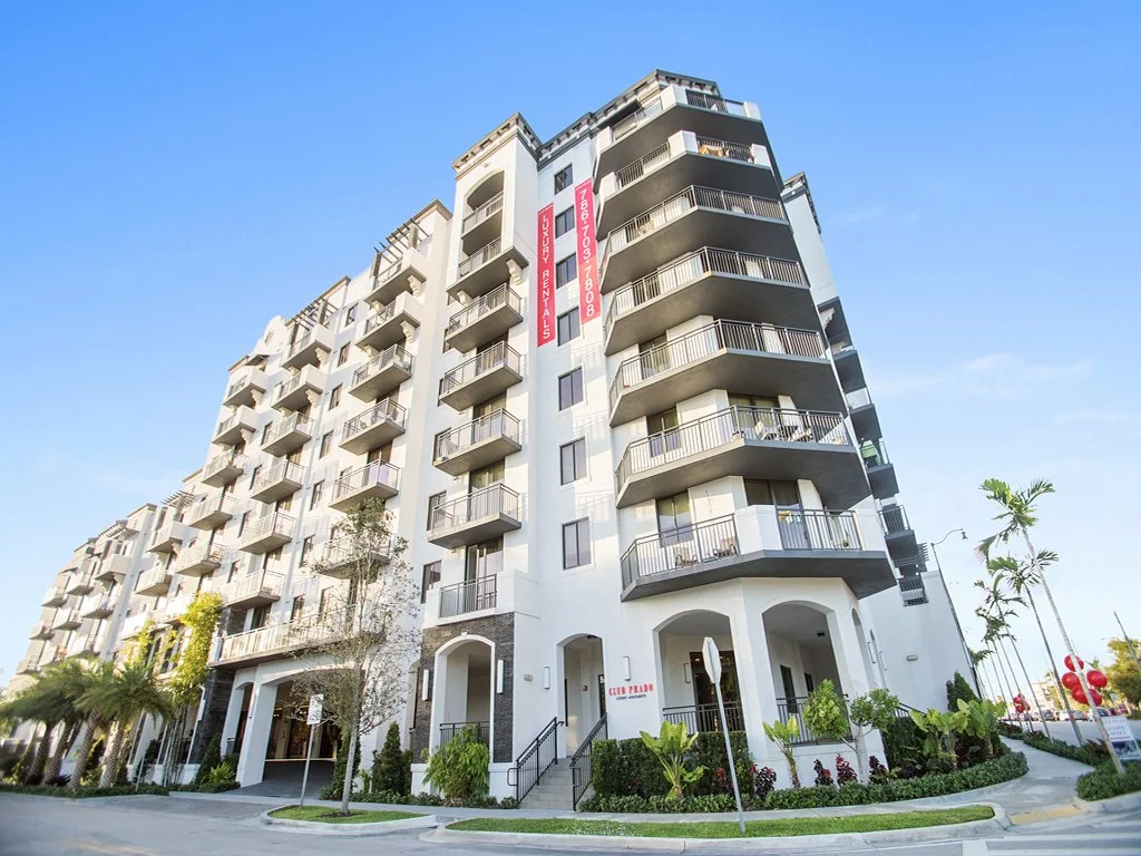A modern multi-story apartment building with balconies, surrounded by landscaping and palm trees, under a clear blue sky.