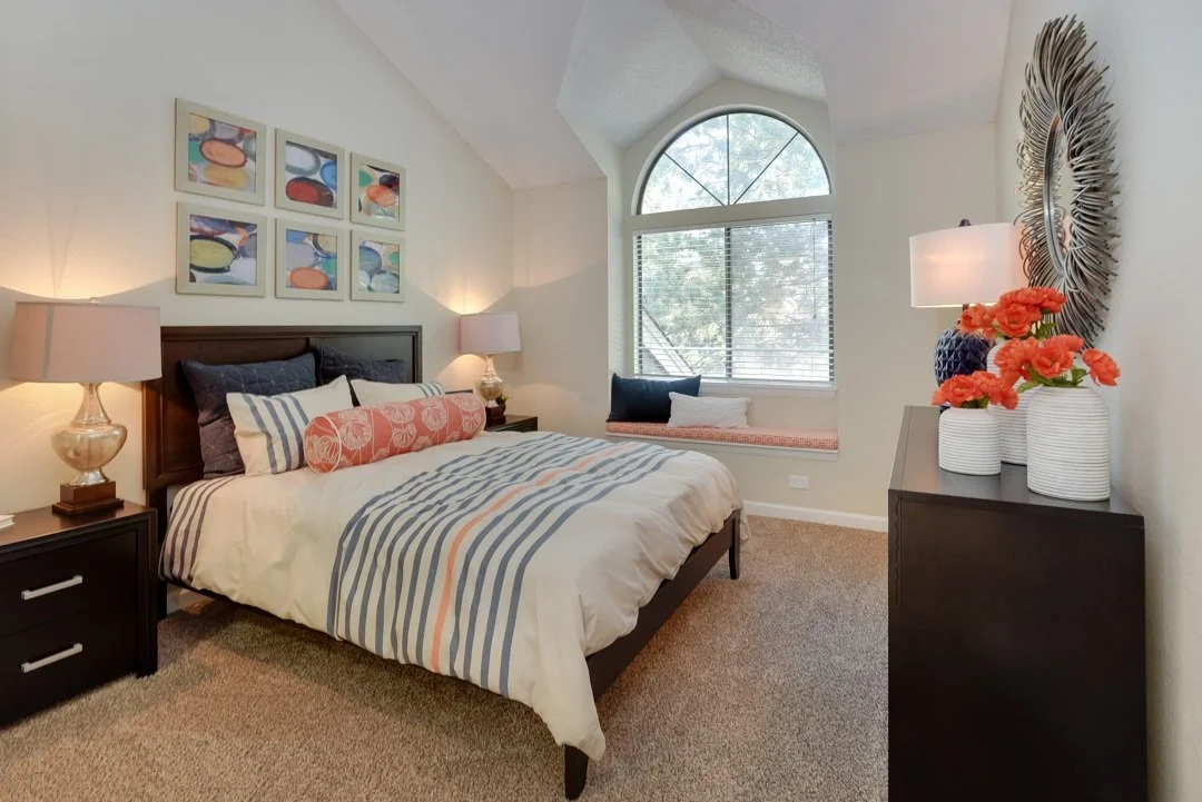 A cozy bedroom with a large window, a bed with striped bedding, and nightstands with lamps. Decor includes wall art and vases with flowers.