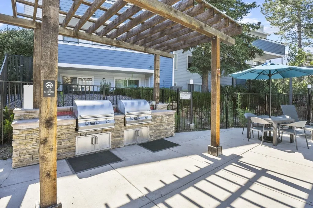 Outdoor communal grilling area with two stainless steel grills under a wooden pergola, patio table with four chairs and a blue umbrella, and a black metal fence in the background.