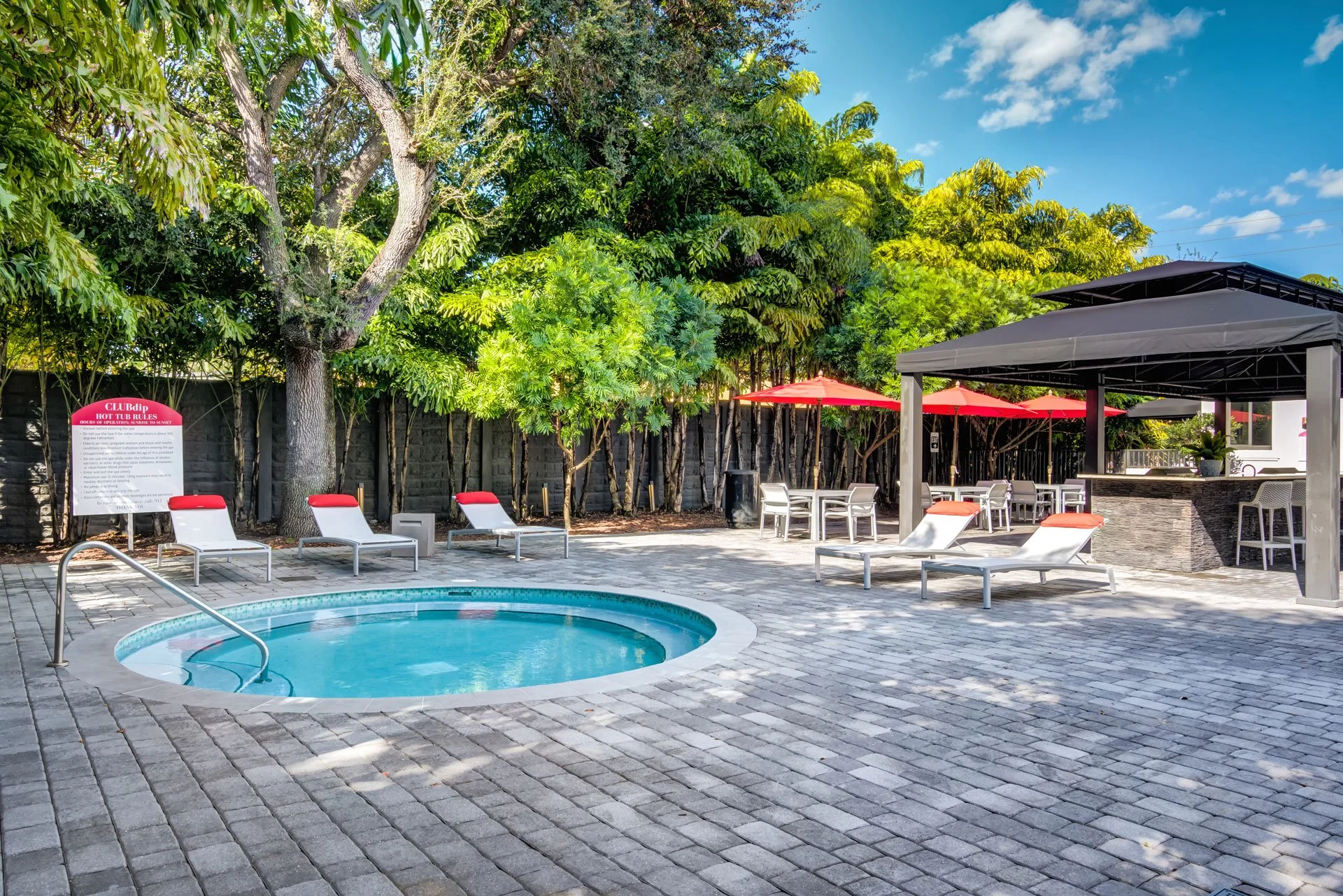 Empty outdoor pool area with lounge chairs, umbrellas, trees, and a shaded bar area.
