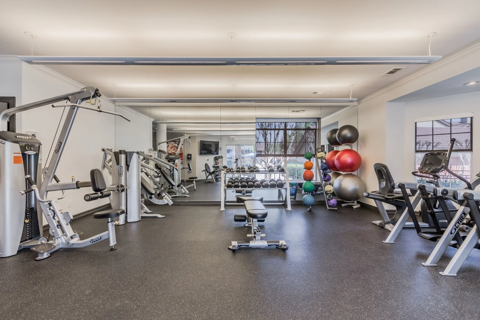 A gym with various exercise equipment, free weights, and balance balls, with large windows providing natural light.