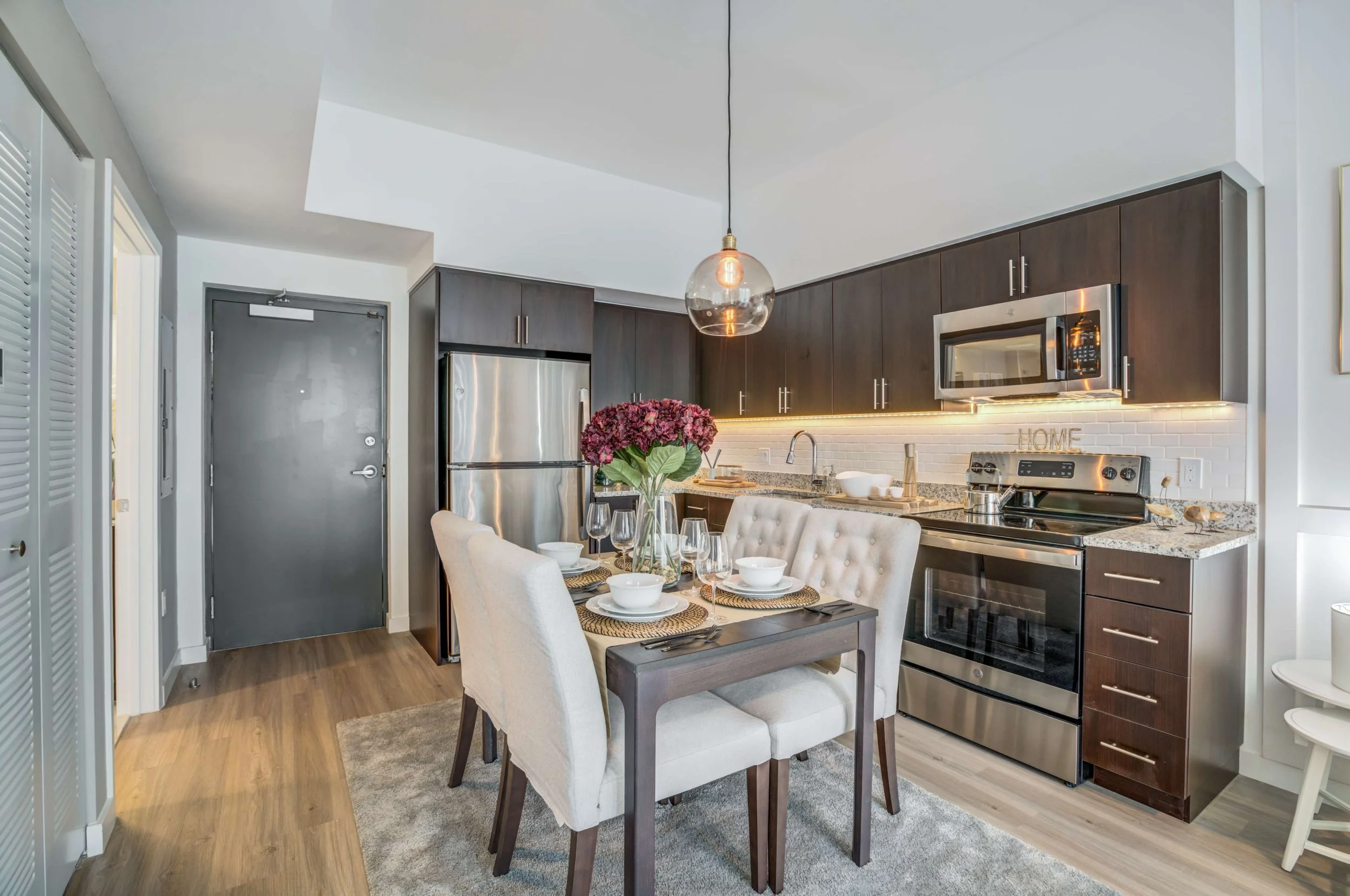 Modern kitchen and dining area with dark wood cabinets, stainless steel appliances, granite countertops, a small dining table with six upholstered chairs, a glass pendant light, and a floral centerpiece.