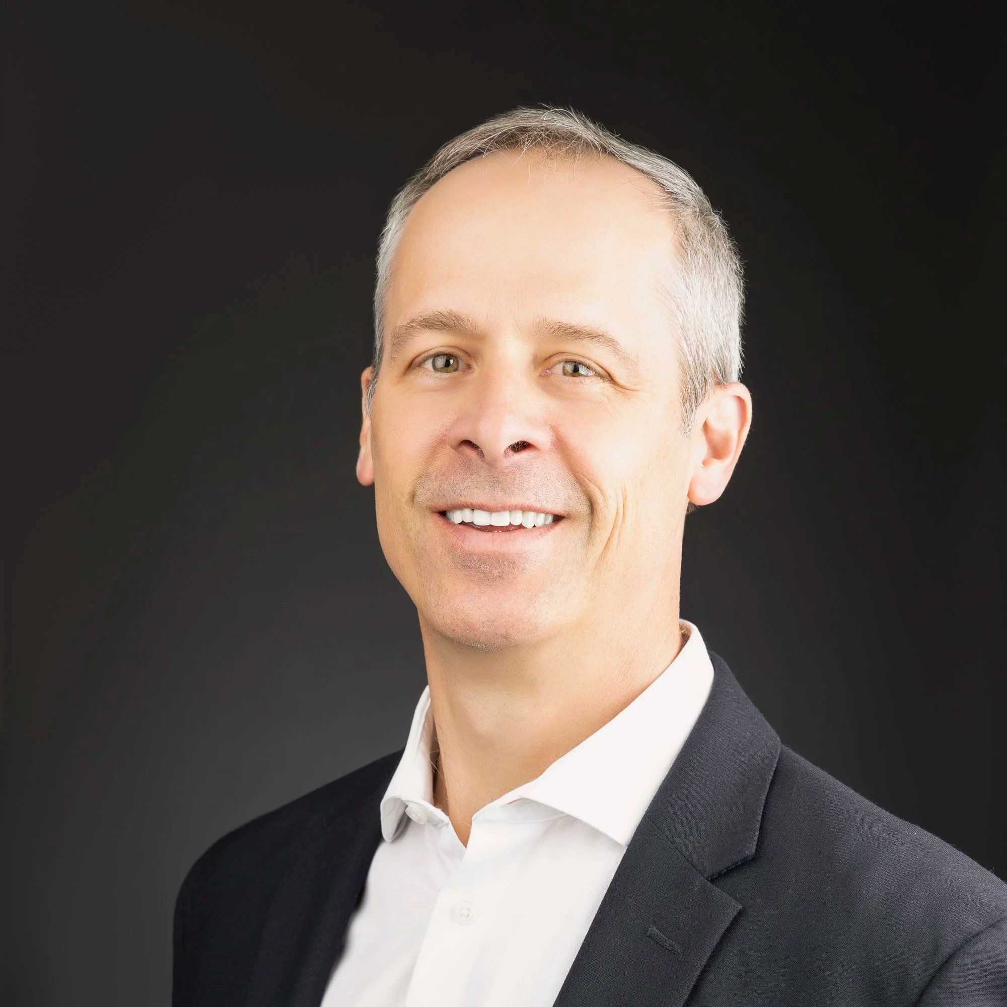 Headshot of a middle-aged man with short gray hair and blue eyes, wearing a white shirt and dark blazer, smiling against a dark background.
