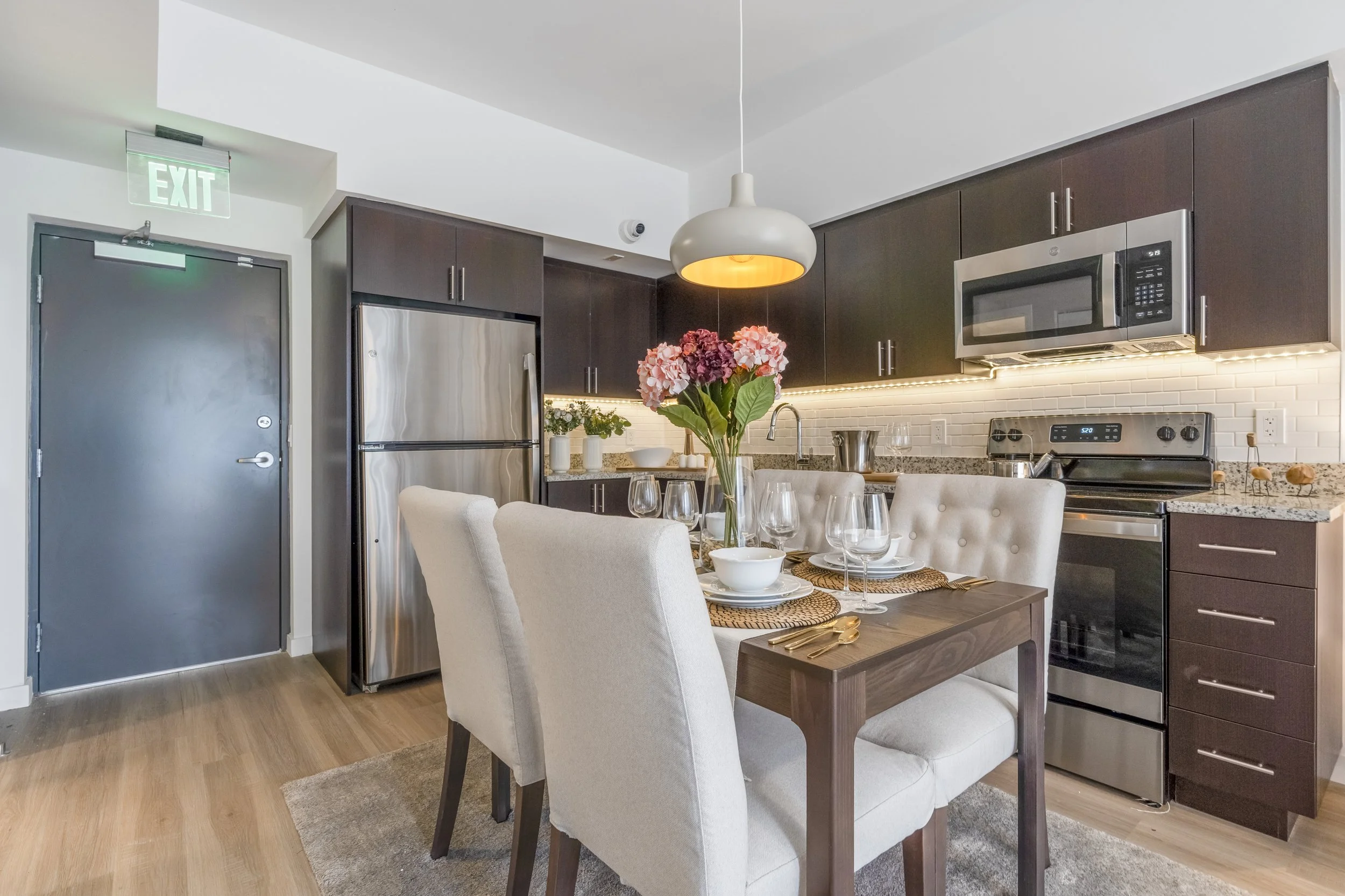 Modern kitchen with dark upper cabinets, stainless steel appliances, and a dining table set with white upholstered chairs, a vase of pink and purple flowers, and tableware.
