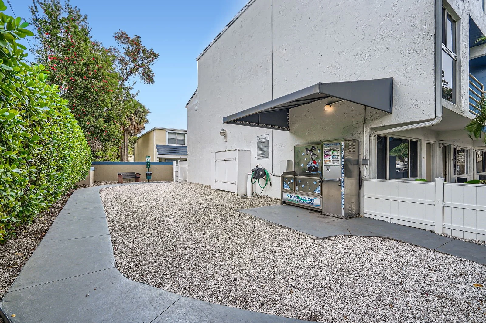 Side of a white building with a gravel pathway, some green shrubbery, and trees. There is a black vending machine under a small awning, and a hose reel nearby.