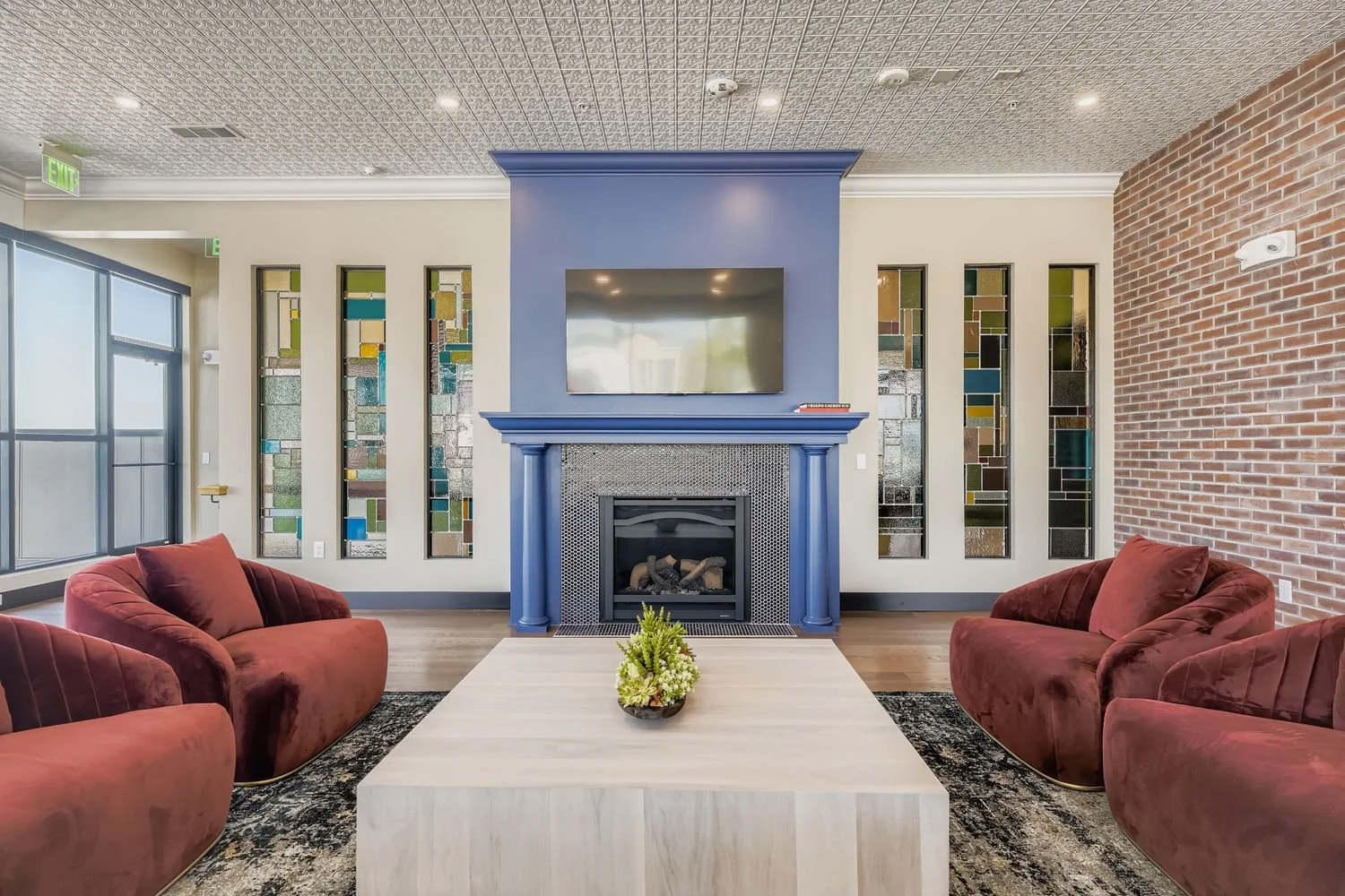 Living room with red velvet sofas, a central white coffee table with a plant, a blue fireplace with a mounted TV, stained glass windows on the walls, large windows on the left, and a brick accent wall on the right.
