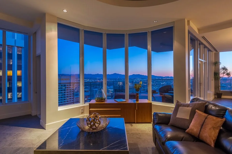 Living room with large curved floor-to-ceiling windows showing a city view at dusk, with mountains in the background, modern furniture, and houseplants.