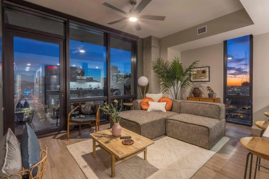 Modern living room with large floor-to-ceiling windows showing a cityscape during sunset, a sectional sofa, wooden coffee table, potted plants, and decorative items.