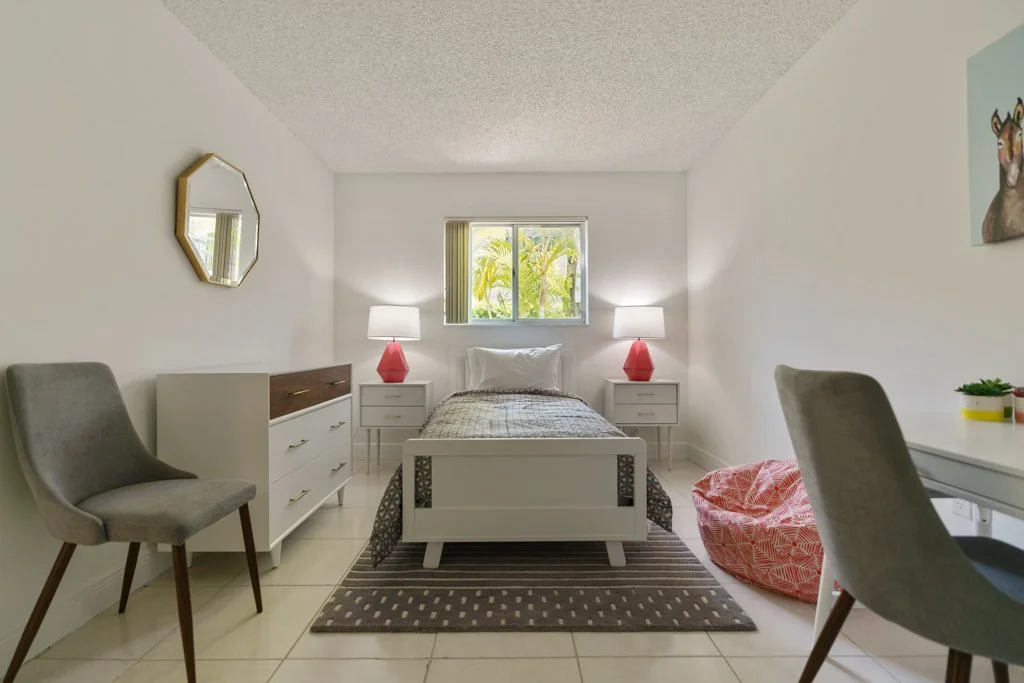A bedroom with a white bed, two nightstands with matching red lamps, a small window with greenery outside, a gray chair on the left, a bean bag chair on the right, and a striped rug on tiled floor.