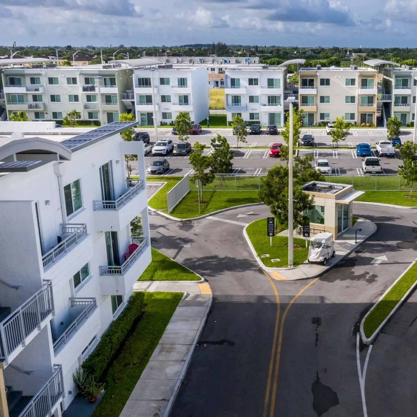 A view of a modern apartment complex with multiple white buildings, green lawns, parking lots with several cars, and a street with a small golf cart.