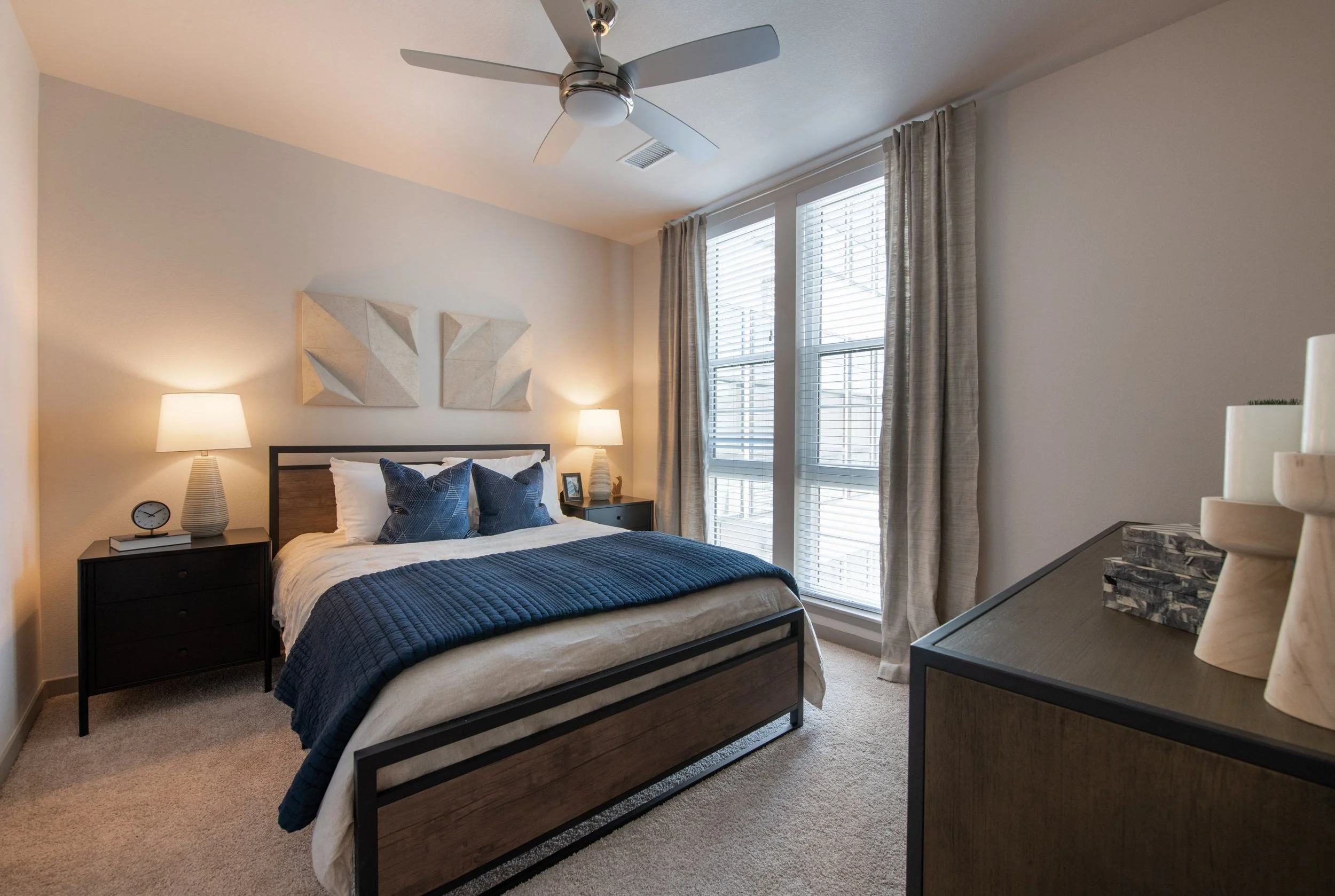 A modern bedroom with a queen-sized bed, dark wood frame, white bedding, and blue pillows. Two matching lamps on nightstands flank the bed. Large windows with blinds and curtains allow natural light. Minimalist wall art hangs above the headboard, and